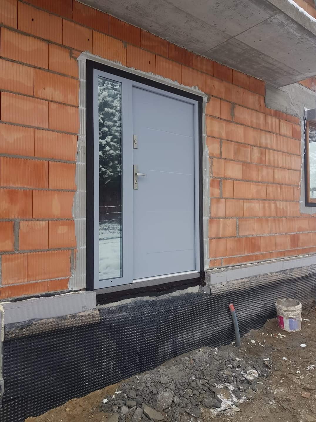Szare drzwi zewnętrzne z szybą boczną, zamontowane w surowym murze z czerwonej cegły. Widoczne ocieplenie fundamentów i fragment budynku w stanie budowy.