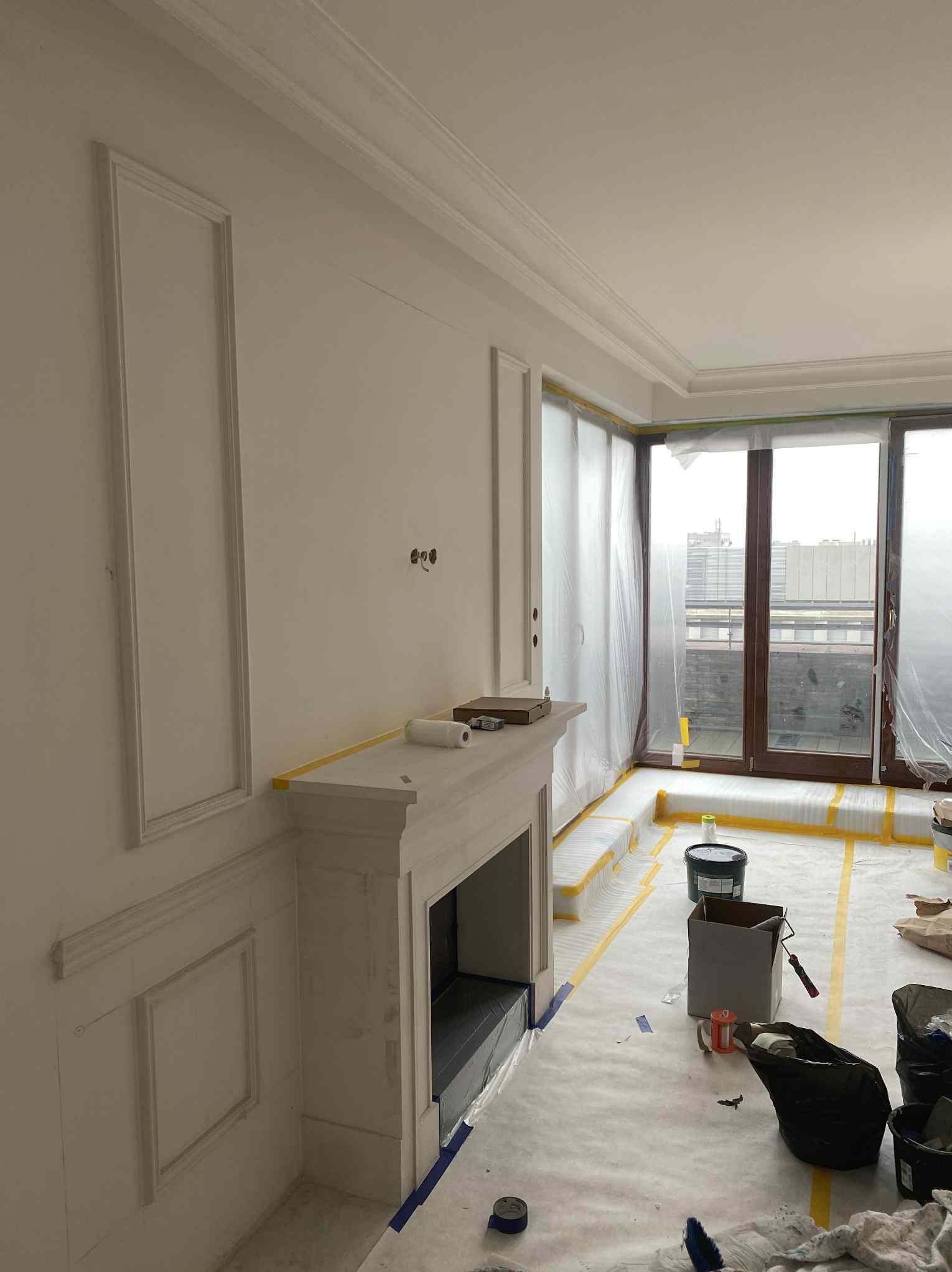 Białe ściany z dekoracyjnymi listwami i kominkiem w trakcie malowania. Zabezpieczone okna i podłoga folią malarską. Widoczne narzędzia i materiały malarskie.