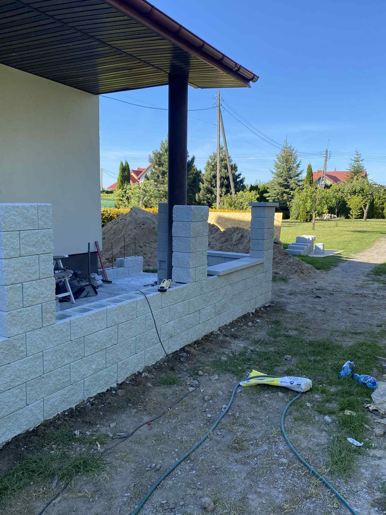 Budowa murku z bloczków betonowych na zewnątrz budynku. Widoczne narzędzia i materiały budowlane. Częściowo ukończona konstrukcja w jasnej kolorystyce.