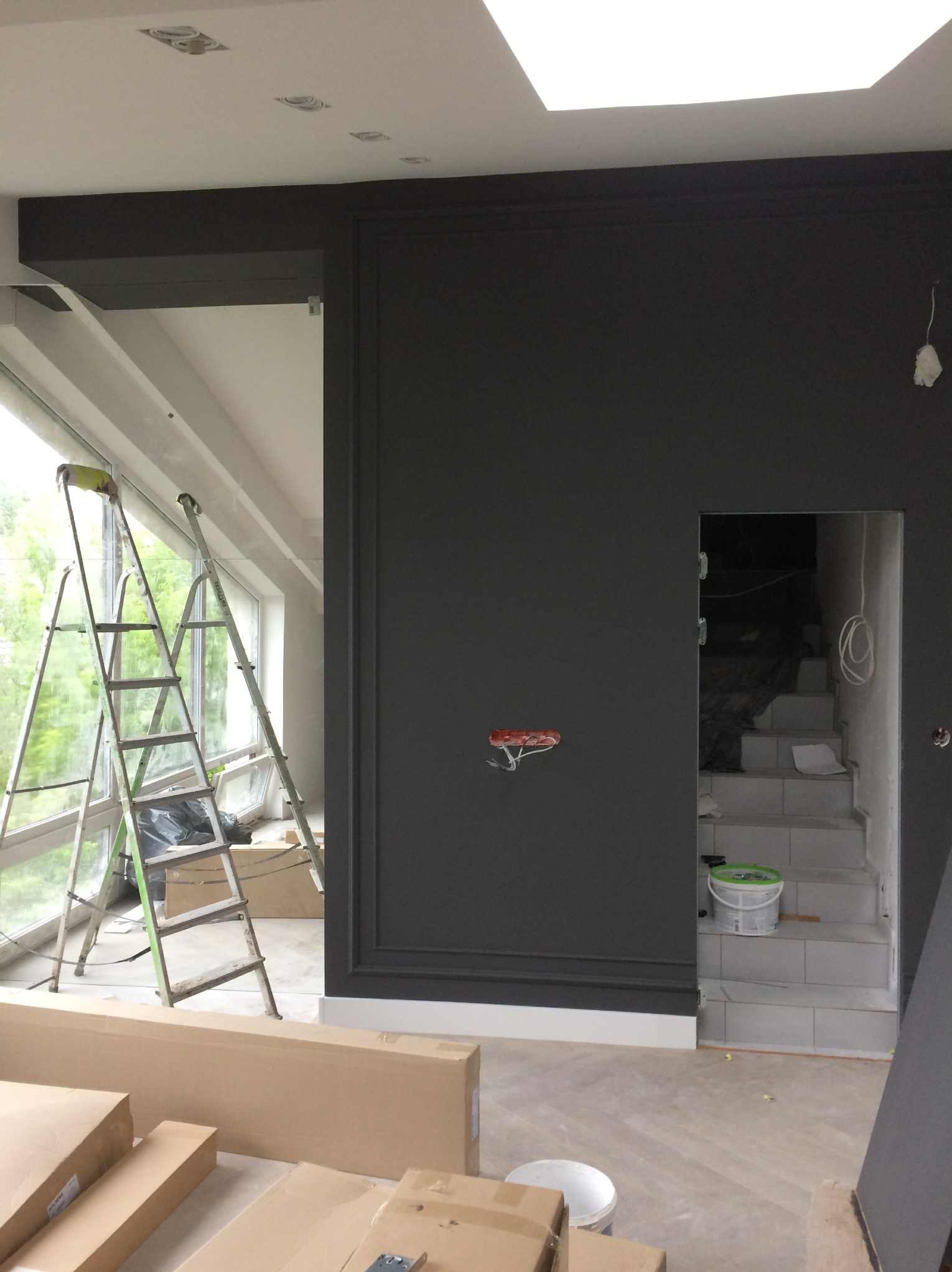 Wnętrze w trakcie malowania na ciemny kolor. Widoczne drabiny, kartony, puszki z farbą i fragment schodów. Ściana z dekoracyjną sztukaterią.