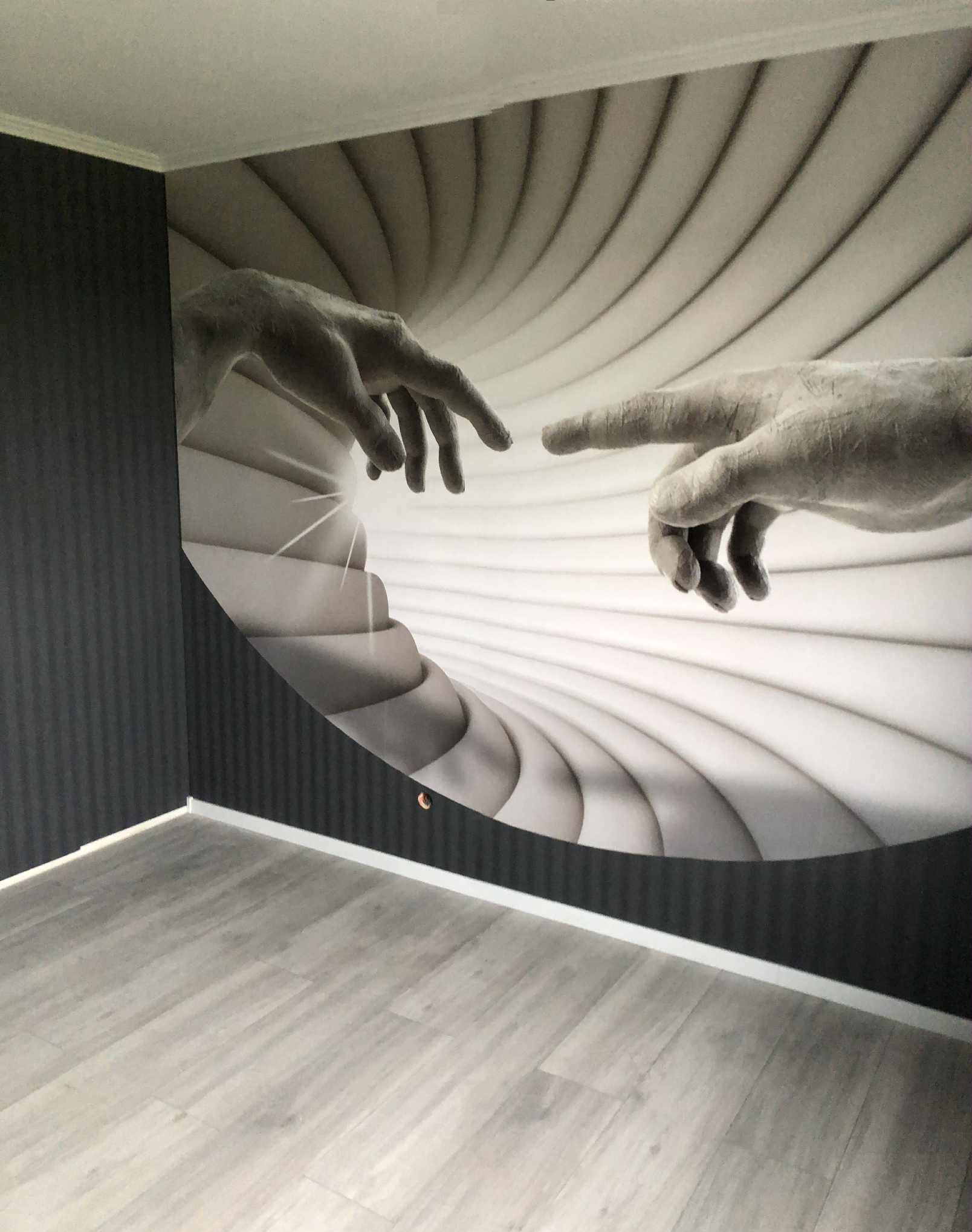 Pokój po remoncie z fototapetą na ścianie przedstawiającą dłonie w stylu 'Stworzenia Adama'. Szare panele podłogowe i ciemna ściana w pionowe pasy.