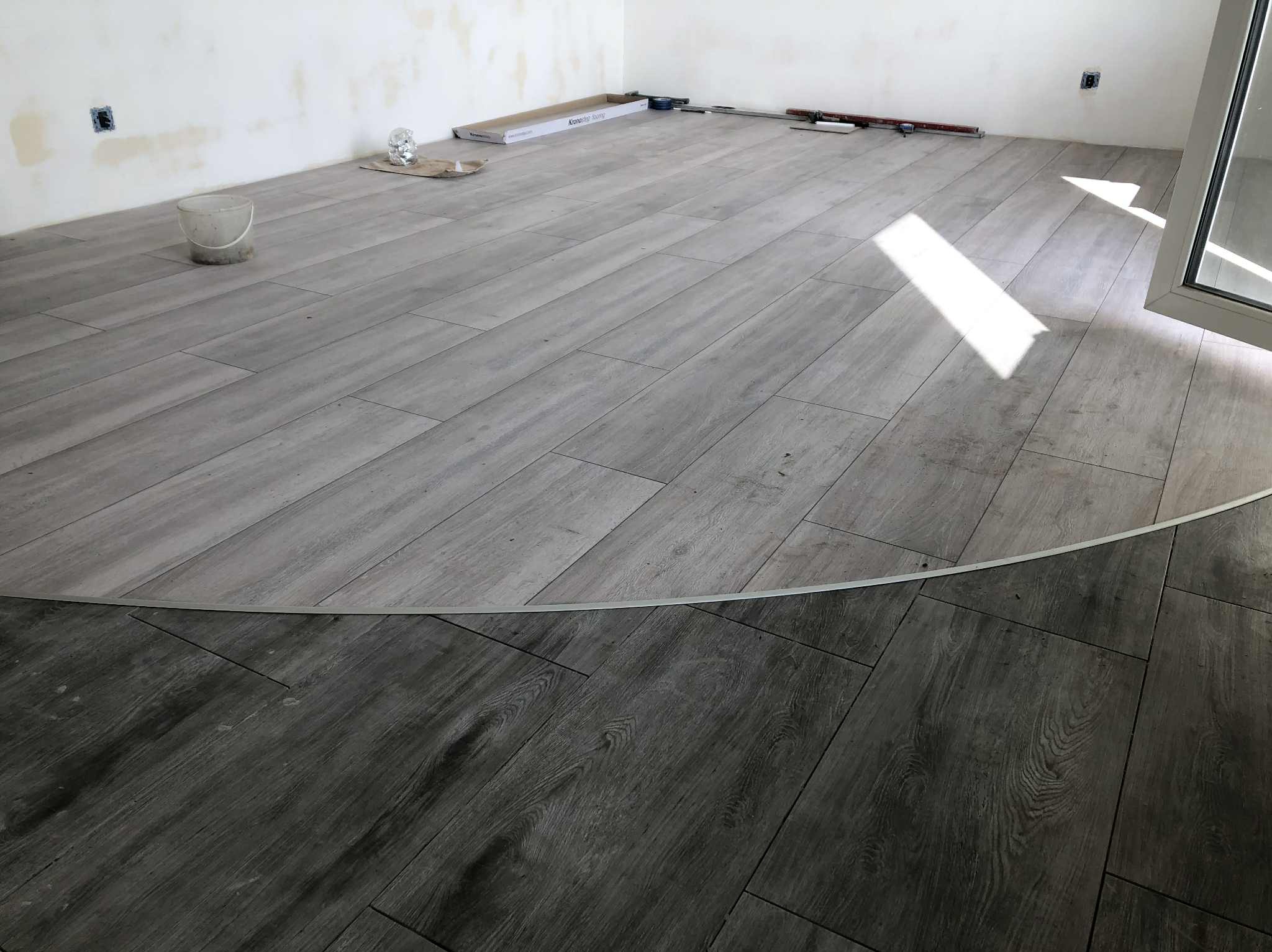 Ukończona podłoga z paneli w odcieniach szarości, z widocznym fragmentem ściany w trakcie wykończenia i narzędziami pracy w tle. Widoczny profil podłogowy.