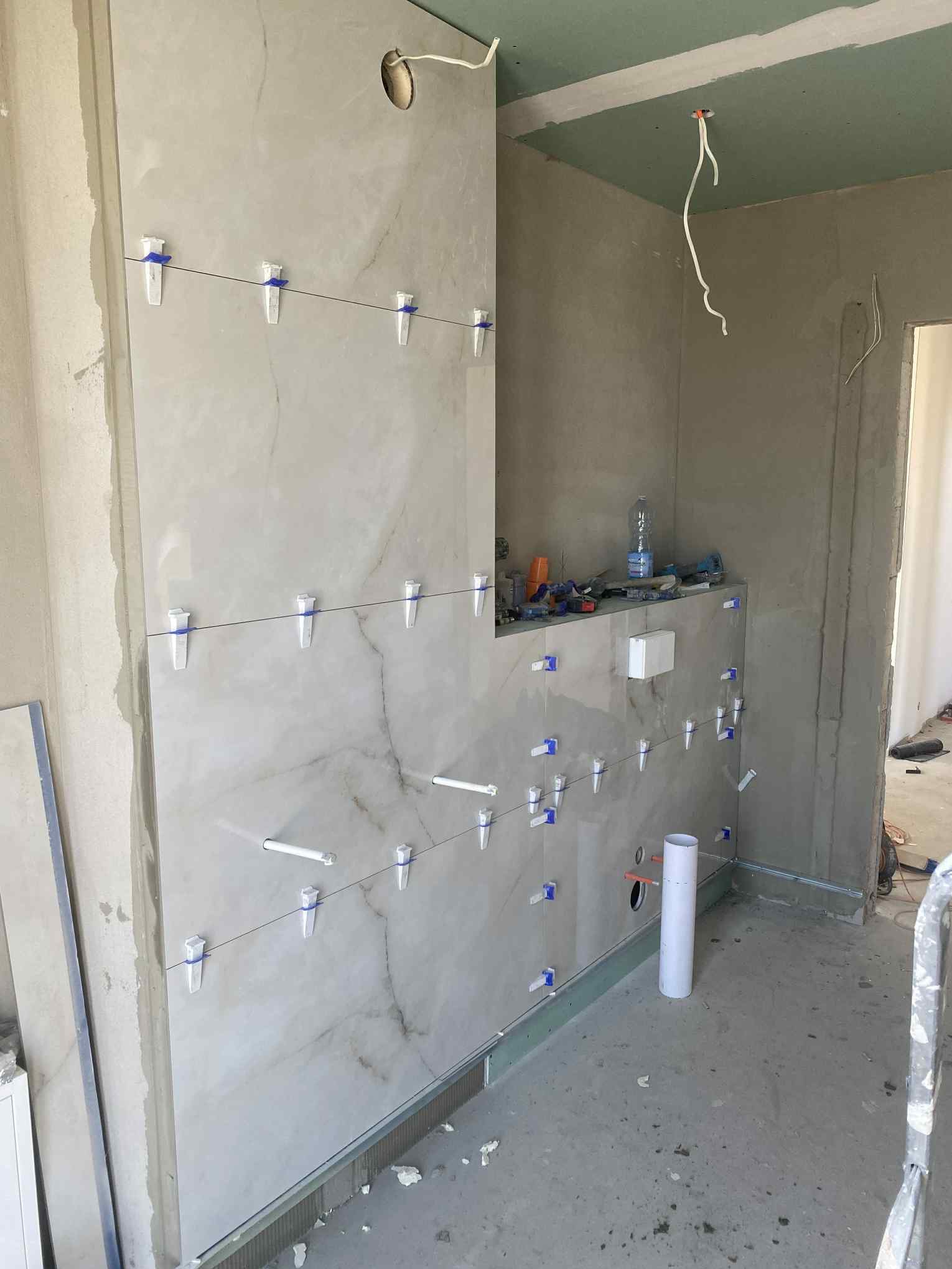 Ściana z marmurowych płytek w trakcie montażu, z użyciem systemu poziomowania. Widoczne narzędzia i elementy instalacji elektrycznej. Sufit z płyt kartonowo-gipsowych.