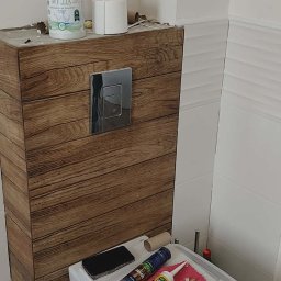 Wykończona obudowa WC z drewnopodobnych paneli, chromowany przycisk spłukiwania. Na sedesie silikony, ściereczka i telefon. W tle białe płytki i podłoga w mozaikę.