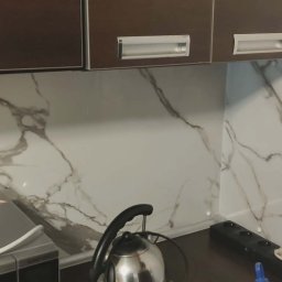 Kuchnia po remoncie: brązowe szafki wiszące nad blatem z marmurowym wzorem. Widoczny czajnik i gniazdko elektryczne. Ukończone wykończenie ściany.