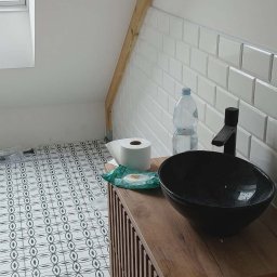 JOS-KOR ANDRZEJ KORCZYŃSKI - Ukończona łazienka na poddaszu w Mińsku Mazowieckim: wzorzysta podłoga, białe płytki metro, drewniana szafka z czarną umywalką. Widoczny fragment okna dachowego.