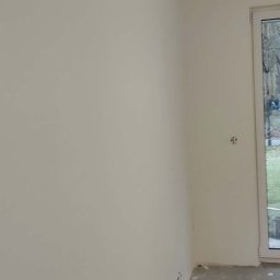 JOS-KOR ANDRZEJ KORCZYŃSKI - Świeżo pomalowane na biało ściany w pokoju z widocznymi gniazdkami elektrycznymi i surową podłogą. Widok na okno z zielenią na zewnątrz.