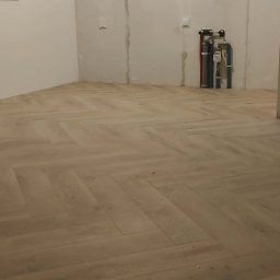 JOS-KOR ANDRZEJ KORCZYŃSKI - Jasny parkiet ułożony we wzór jodełki francuskiej w pustym pokoju. Widoczne rury grzewcze przy ścianie i fragment osoby wchodzącej do pomieszczenia.