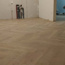 JOS-KOR ANDRZEJ KORCZYŃSKI - Ułożony parkiet w jodełkę w jasnym odcieniu drewna, widoczne rury instalacji na ścianie i fragment osoby podczas prac wykończeniowych w tle.