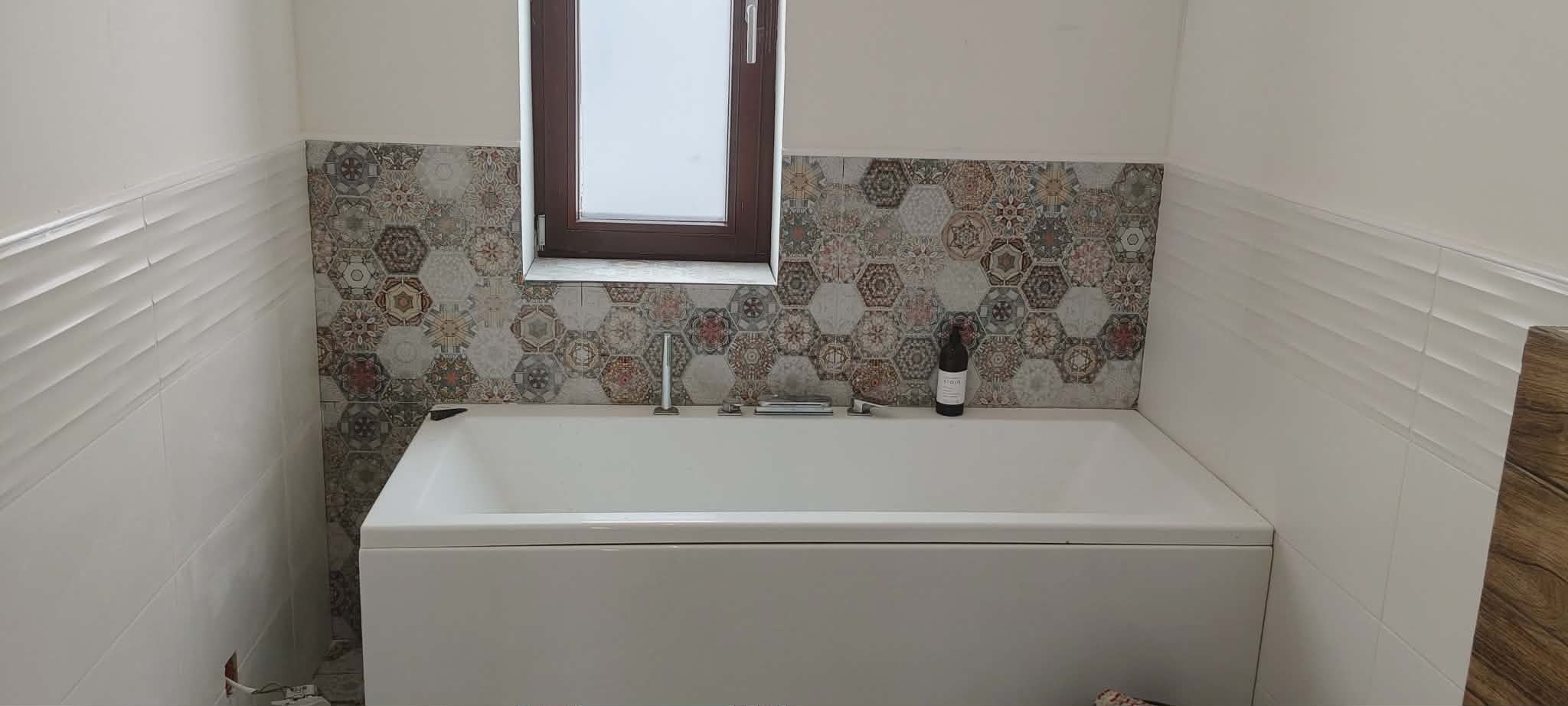 Biała wanna z baterią pod oknem, nad nią dekoracyjne, heksagonalne płytki. Po bokach białe, poziome płytki z delikatnym wzorem. Butelka wina na wannie.