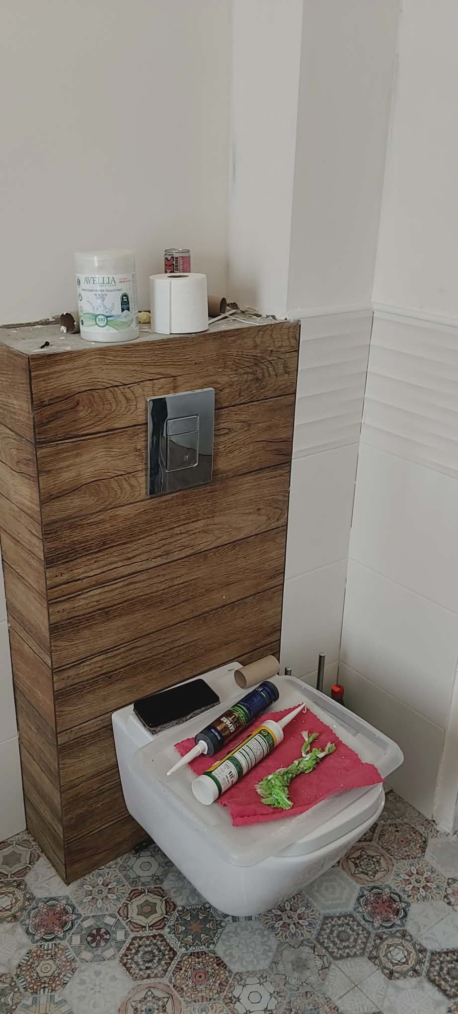 Wykończona obudowa WC z drewnopodobnych paneli, chromowany przycisk spłukiwania. Na sedesie silikony, ściereczka i telefon. W tle białe płytki i podłoga w mozaikę.