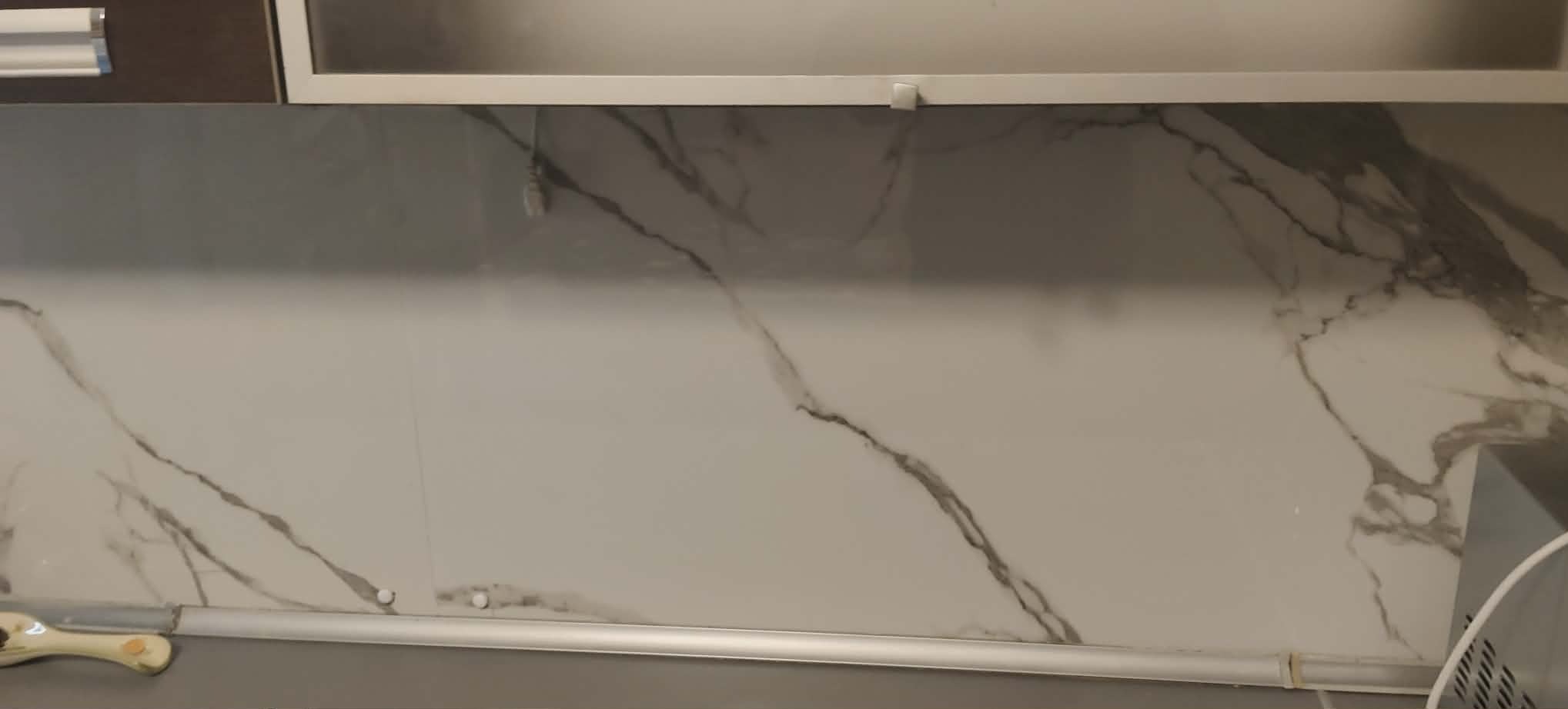 Biała ściana w kuchni z szarym marmurkowym wzorem, widoczne szafki wiszące i listwa przypodłogowa. Minimalistyczny design.