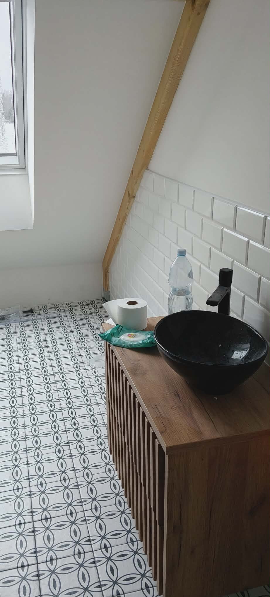 Ukończona łazienka na poddaszu w Mińsku Mazowieckim: wzorzysta podłoga, białe płytki metro, drewniana szafka z czarną umywalką. Widoczny fragment okna dachowego.