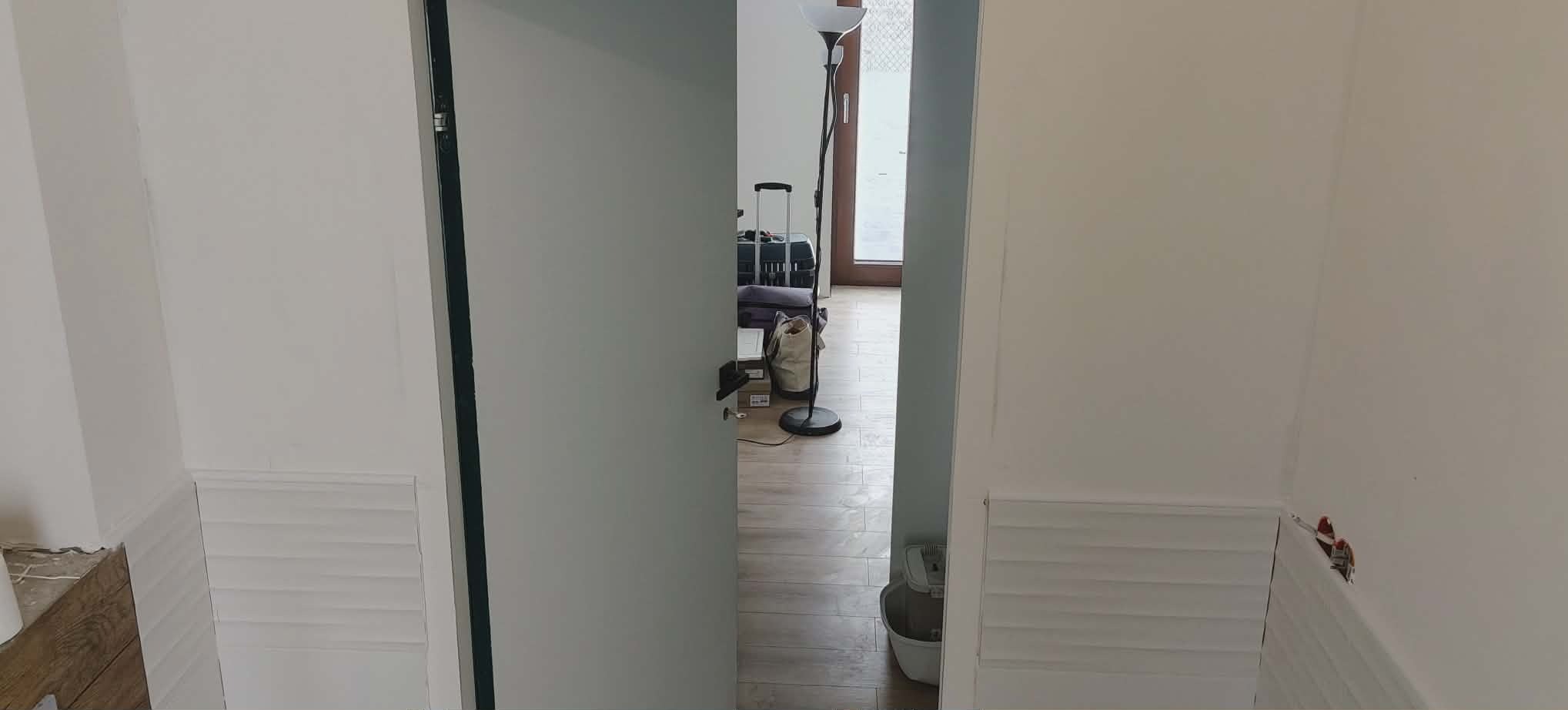 Białe ściany z dekoracyjną listwą przypodłogową i otwarte drzwi do pokoju z drewnianą podłogą, lampą i bagażem. Widok na wykończone wnętrze.