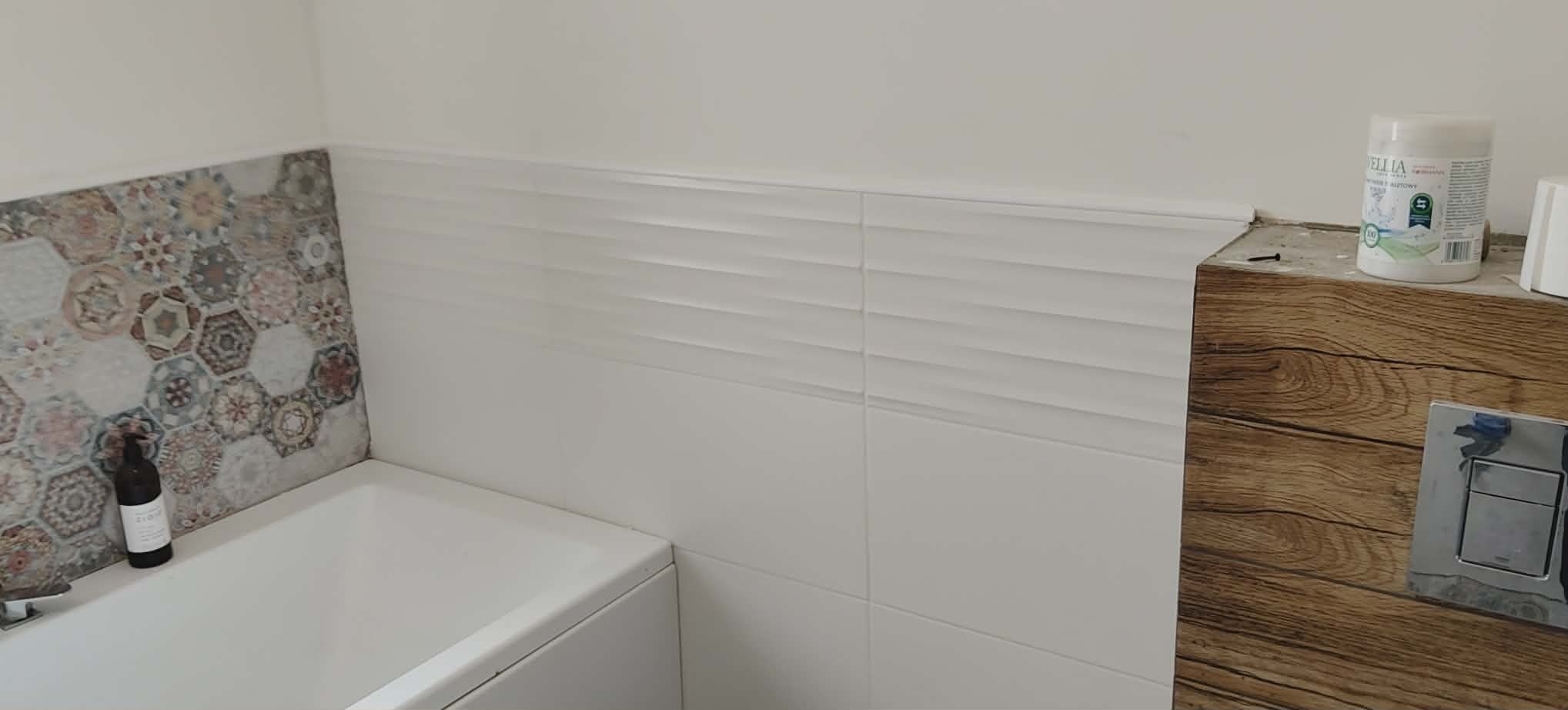 Wykończona łazienka z wanną, białe płytki z poziomymi żłobieniami i dekoracyjna mozaika heksagonalna. Widoczny fragment obudowy WC z drewnopodobną okleiną.