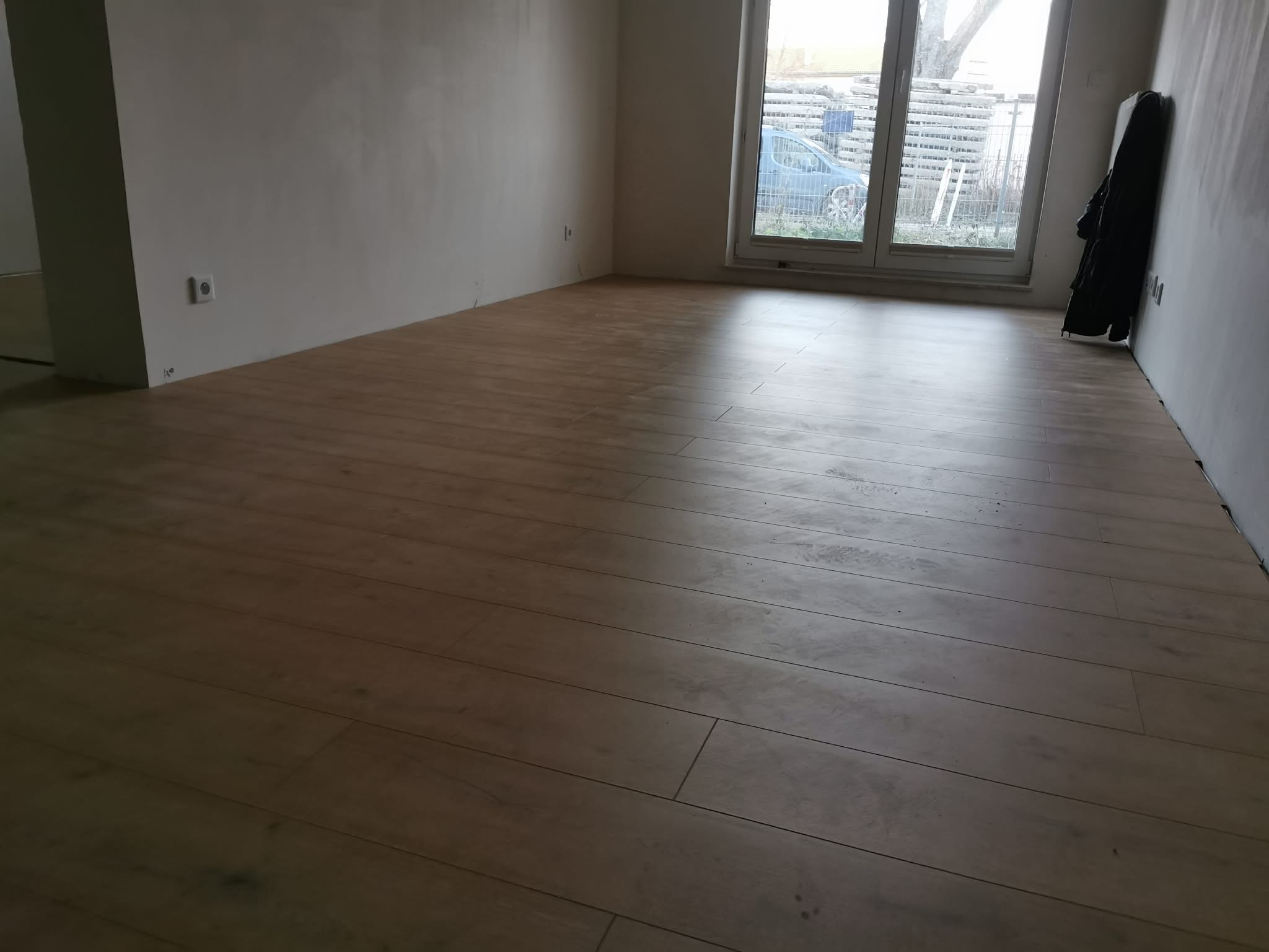 Jasne panele podłogowe ułożone w pustym pokoju z oknem. Ściany w kolorze złamanej bieli, widoczne gniazdka elektryczne. Ubrania wiszą na ścianie.