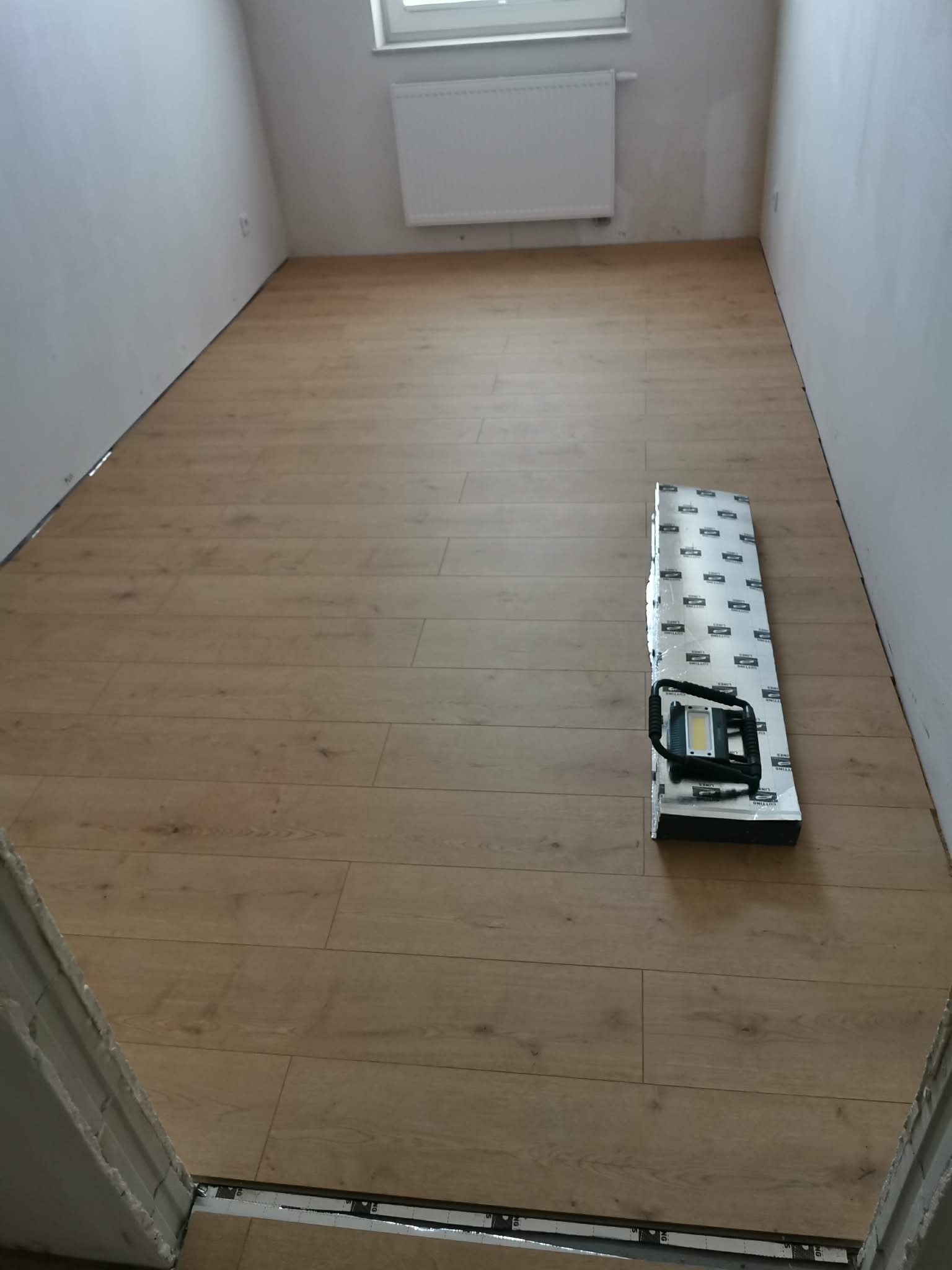Ułożone panele podłogowe w jasnym odcieniu drewna w pokoju z białymi ścianami. Na podłodze leży paczka paneli i lampa robocza. Widoczny fragment progu.
