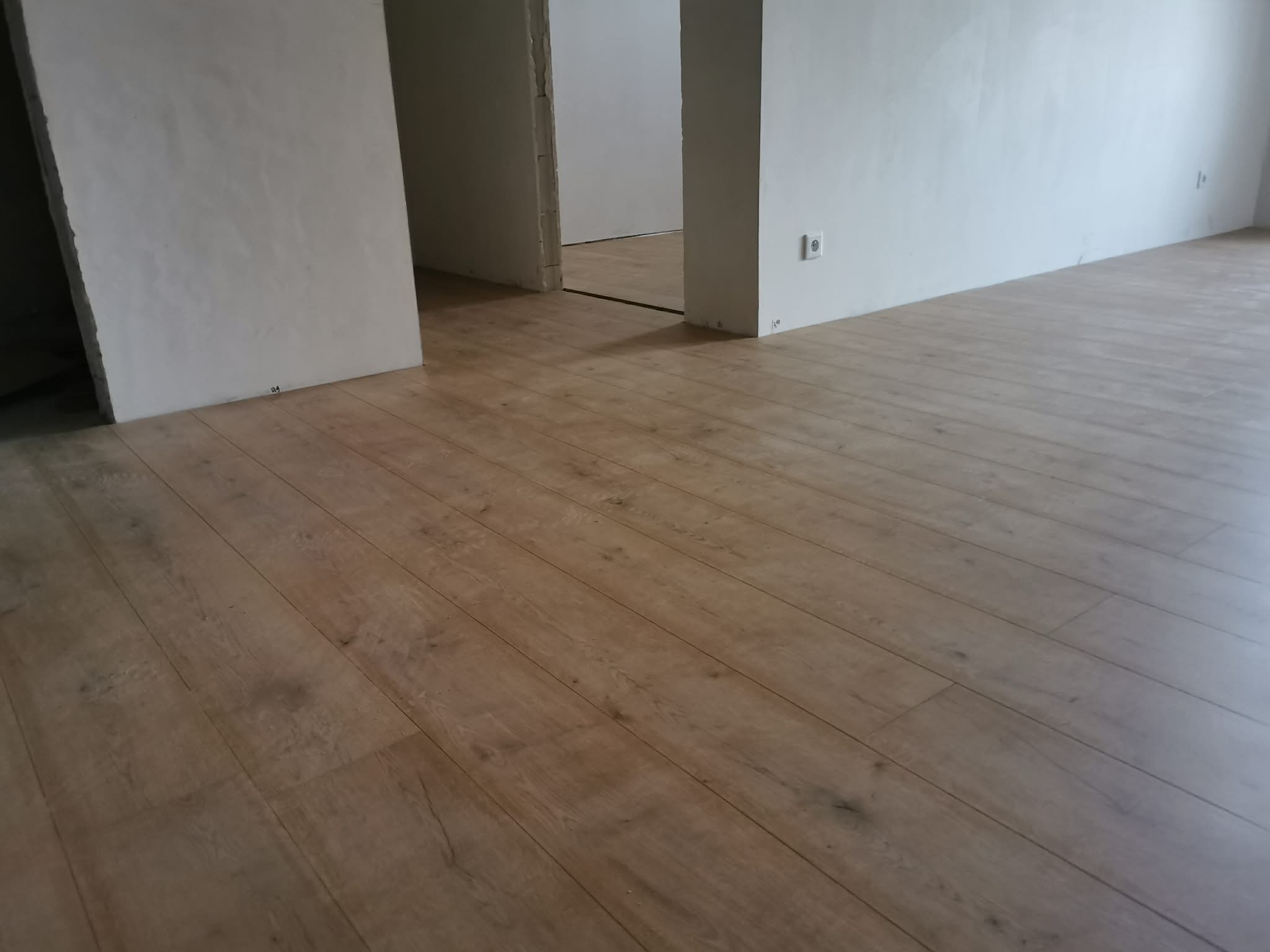 Jasne panele podłogowe ułożone w nowym wnętrzu, widoczne gładkie ściany i gniazdko elektryczne. Perspektywa z niskiego kąta podkreśla wzór drewna.