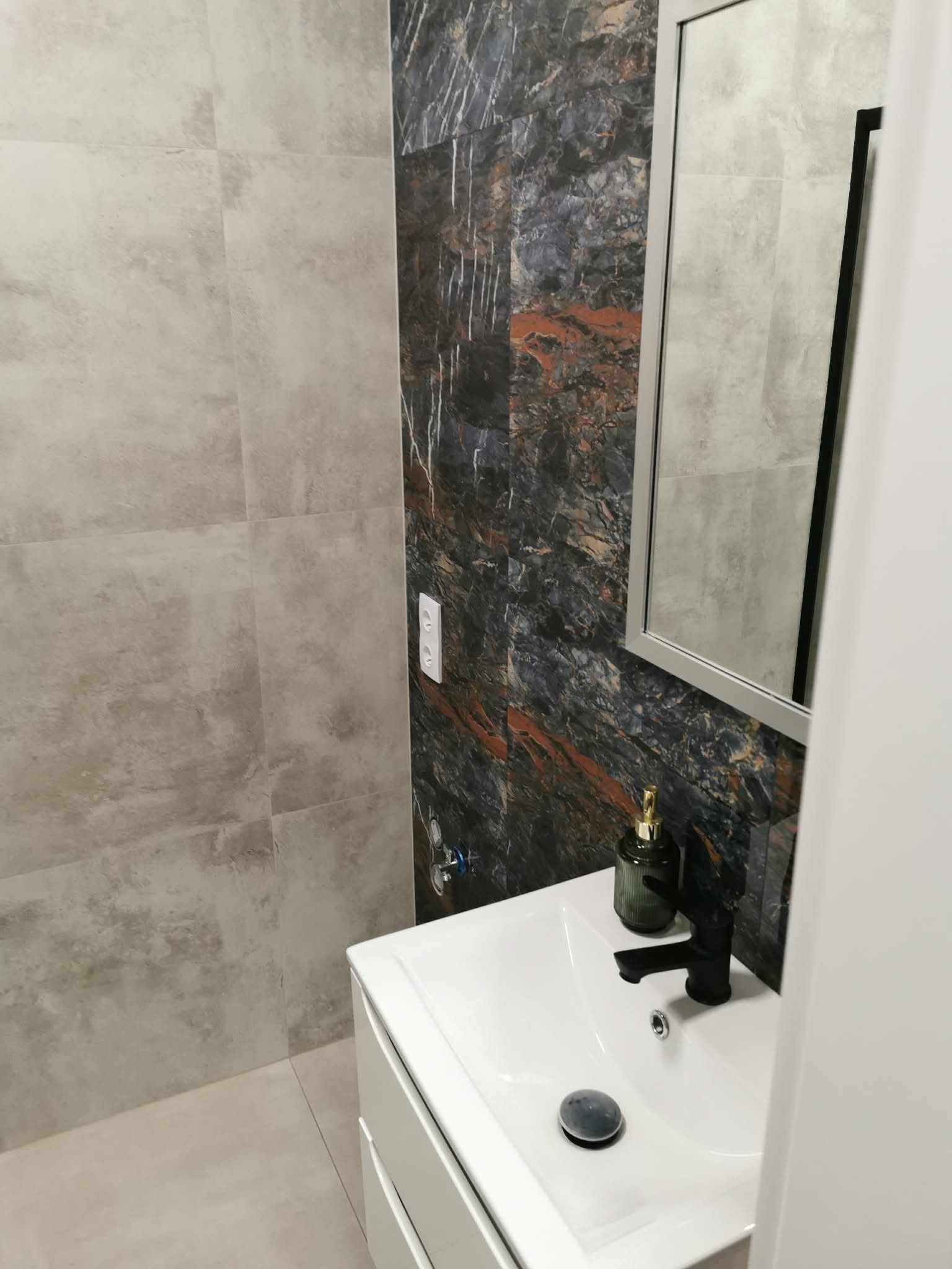 Wykończona łazienka z umywalką, lustrem i płytkami imitującymi marmur. Widoczny fragment ściany z gniazdkiem elektrycznym. Nowoczesny design.