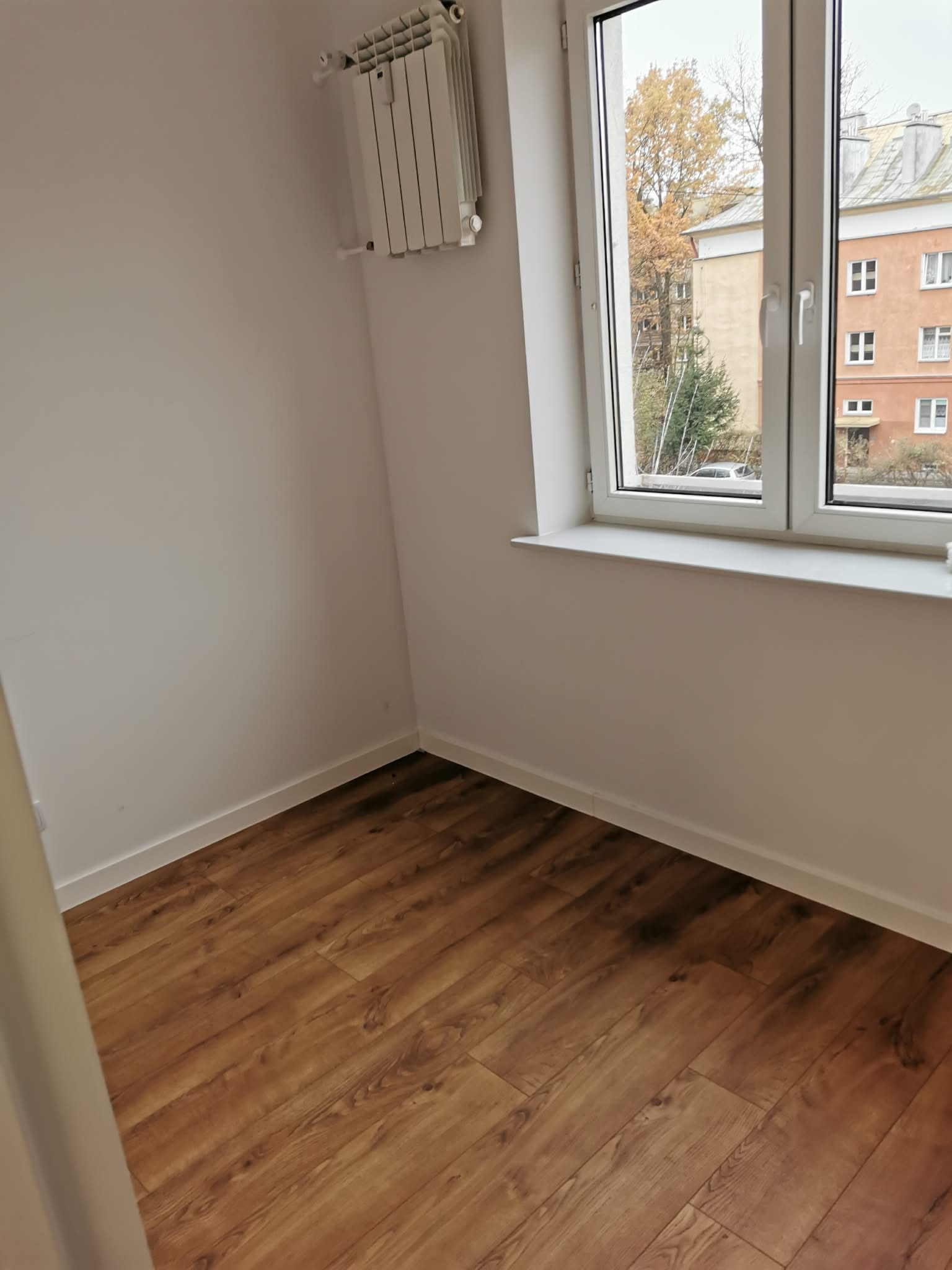 Jasne wnętrze po remoncie: panele podłogowe w odcieniach brązu, białe ściany, okno z widokiem na budynek i drzewa, grzejnik. Minimalistyczny design.