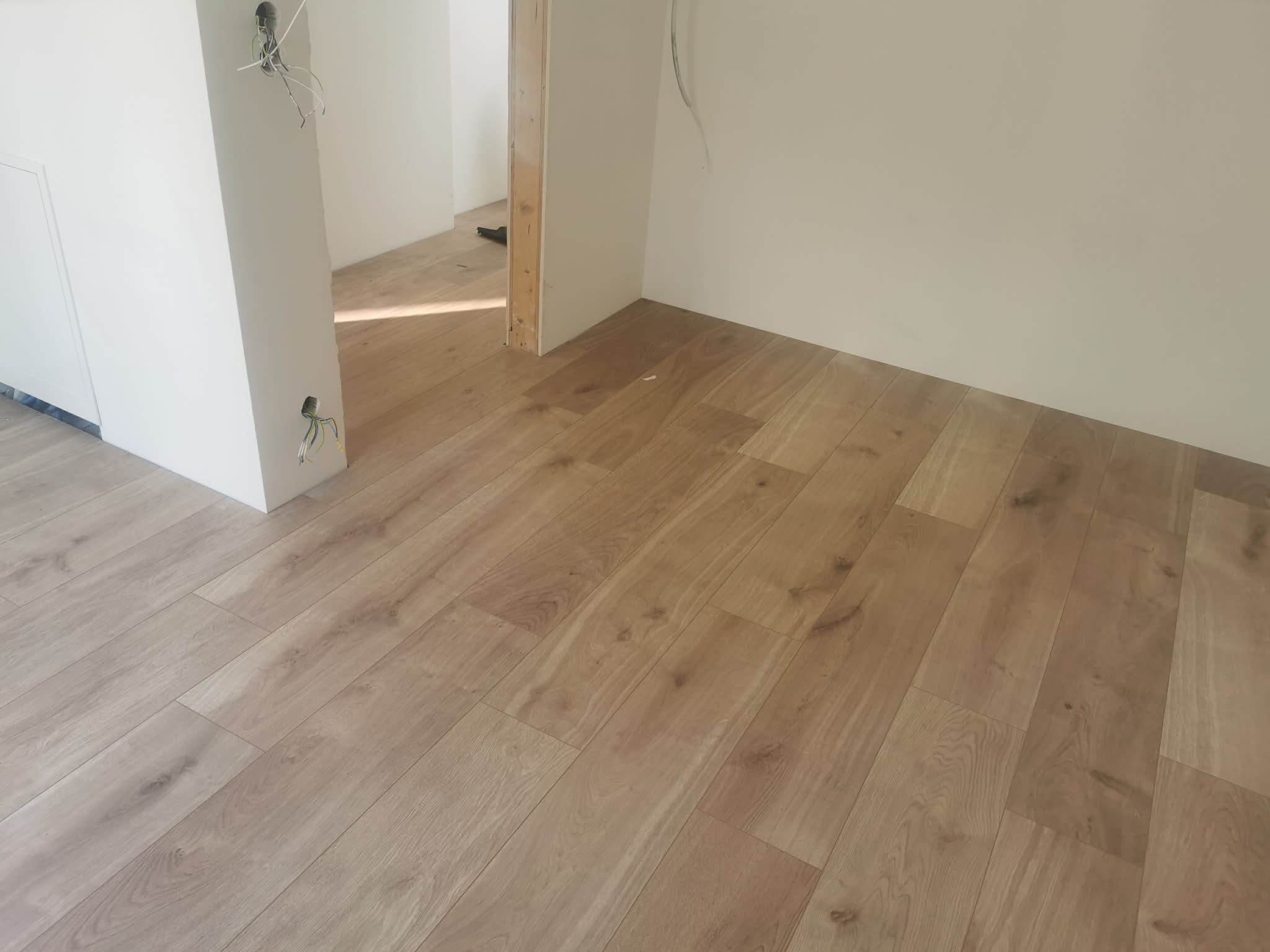 Jasne panele podłogowe ułożone w nowym wnętrzu, widoczne kable elektryczne wystające ze ściany, sugerujące świeżo zakończony etap prac wykończeniowych.