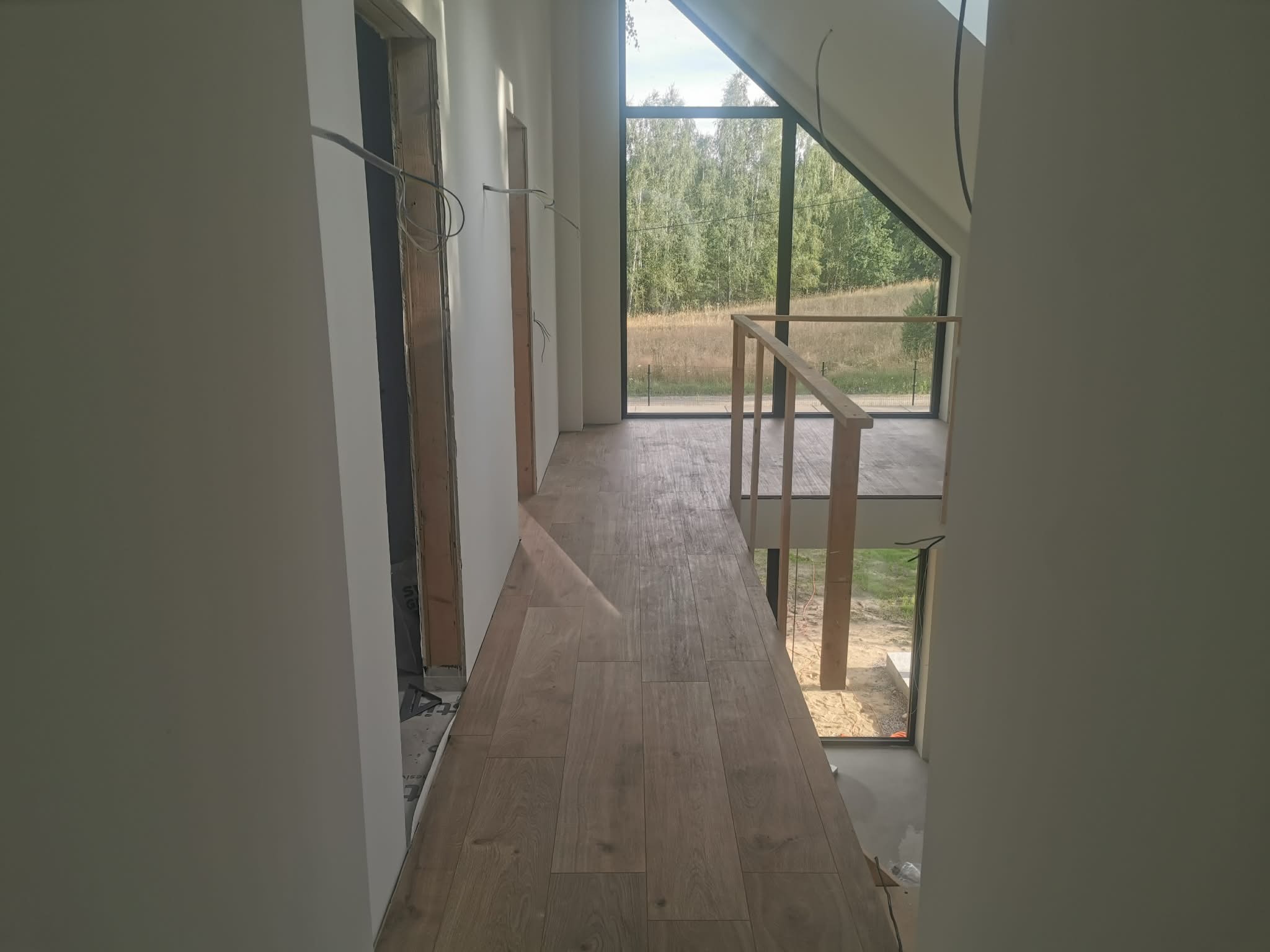 Jasne wnętrze domu w trakcie wykańczania: drewnopodobne panele na podłodze, duże okno z widokiem na zieleń, surowe ościeżnice drzwiowe i wystające przewody.