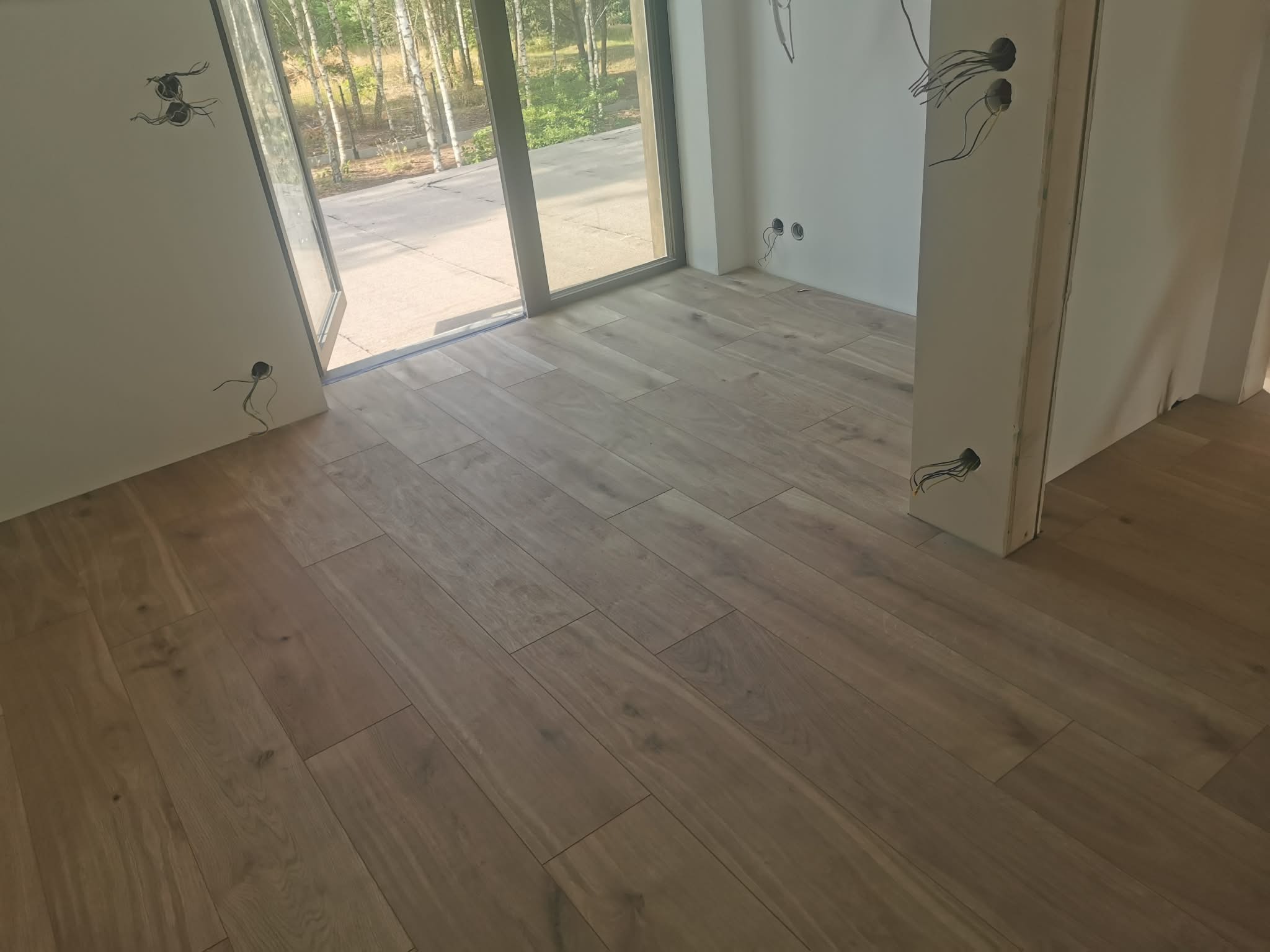 Ułożone panele podłogowe w jasnym odcieniu drewna, widoczne gniazdka elektryczne bez osprzętu i otwarte drzwi balkonowe z widokiem na zieleń.