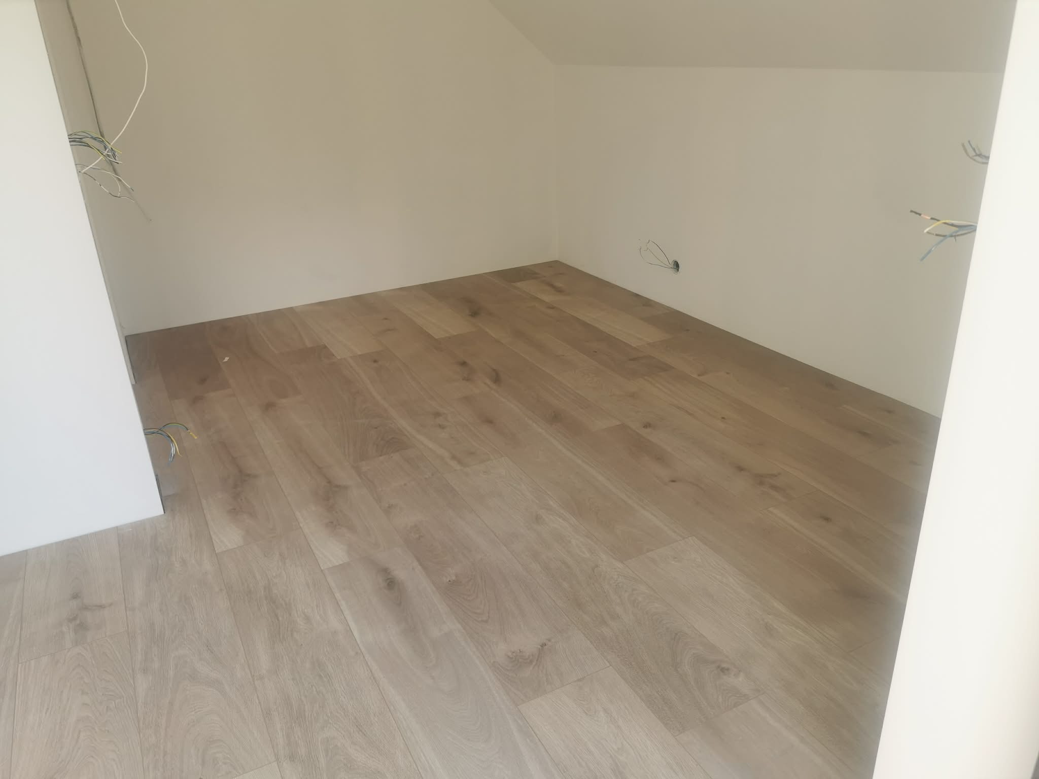 Jasne panele podłogowe ułożone w pokoju na poddaszu, widoczne kable elektryczne wystające ze ściany, świeżo po montażu. Ujęcie z niskiego kąta.
