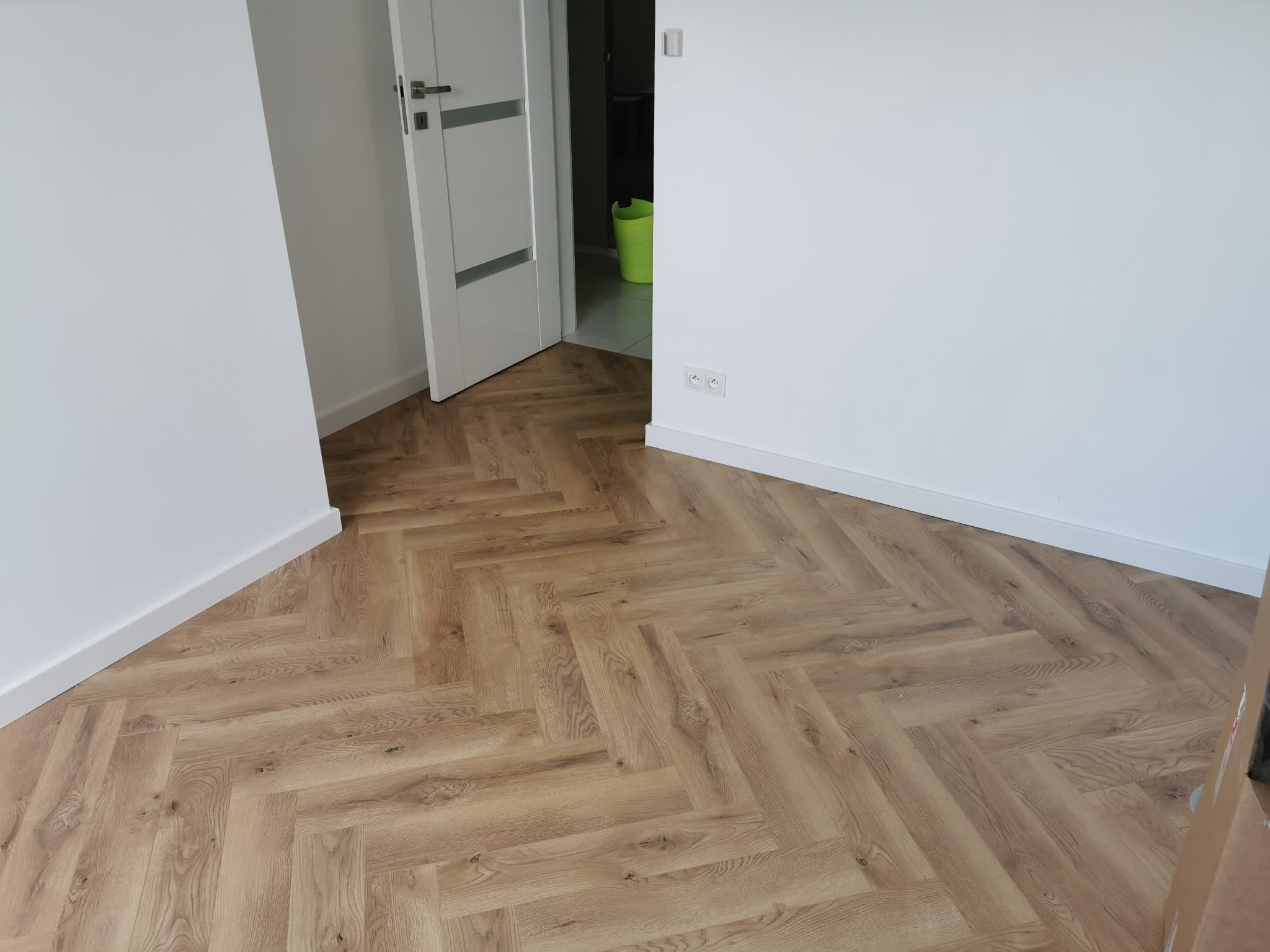 Jasne panele ułożone w jodełkę w pokoju z białymi ścianami i otwartymi, białymi drzwiami. Widoczne gniazdka elektryczne i listwy przypodłogowe.