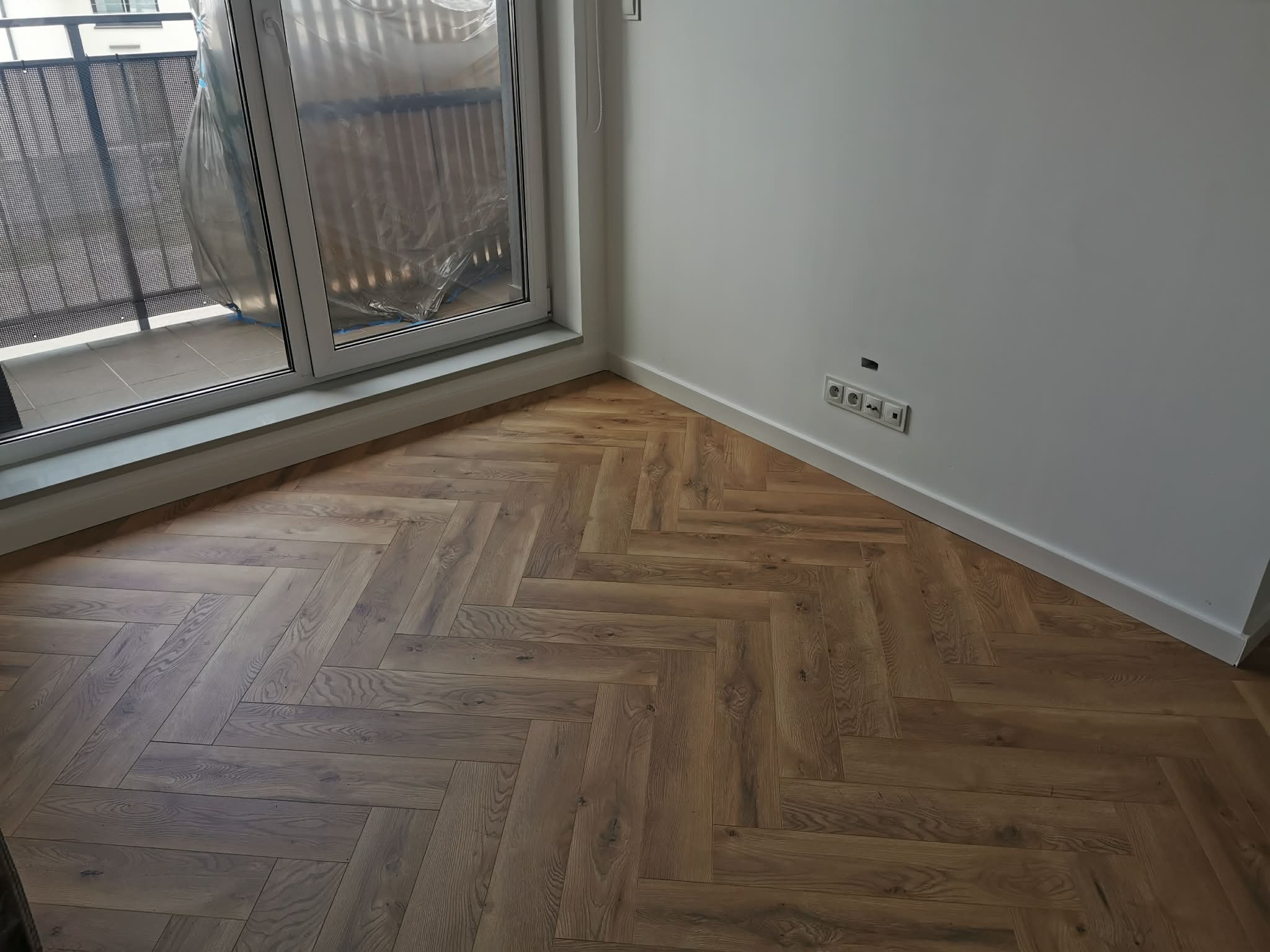 Ukończona podłoga z parkietu ułożonego w jodełkę francuską, widok z góry, przy oknie balkonowym i białych ścianach z gniazdkami elektrycznymi.