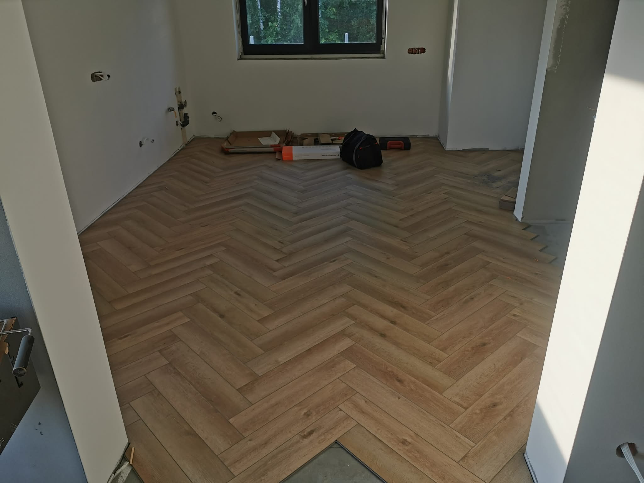 Ułożony parkiet w jodełkę w jasnym pomieszczeniu z oknem. Widoczne narzędzia i materiały budowlane. Częściowo wykończone ściany. Ujęcie z niskiego kąta.