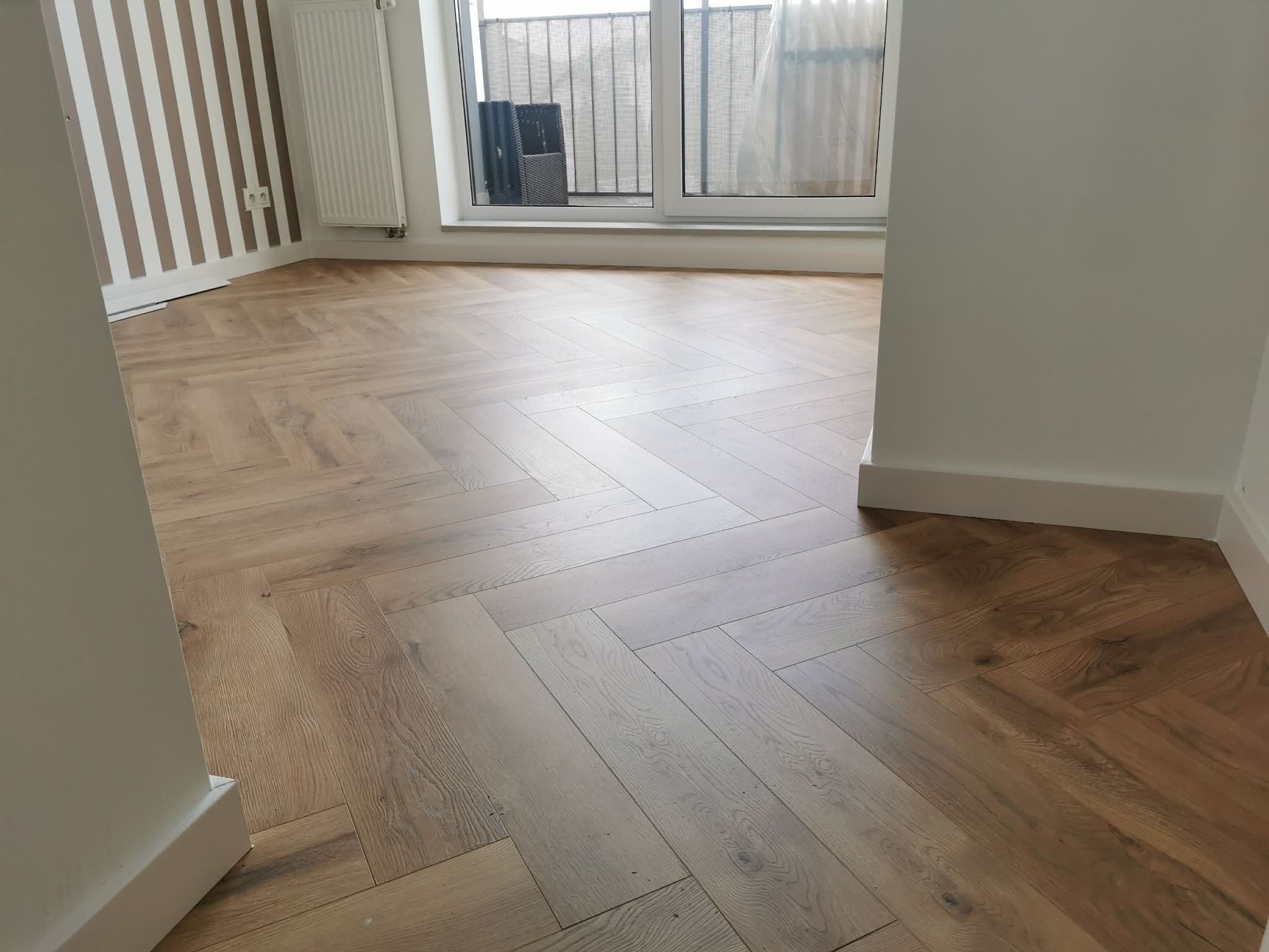 Jasny parkiet ułożony w jodełkę francuską w nowym wnętrzu. Widoczne listwy przypodłogowe i fragment ściany w pionowe pasy. Okno z widokiem na balkon.