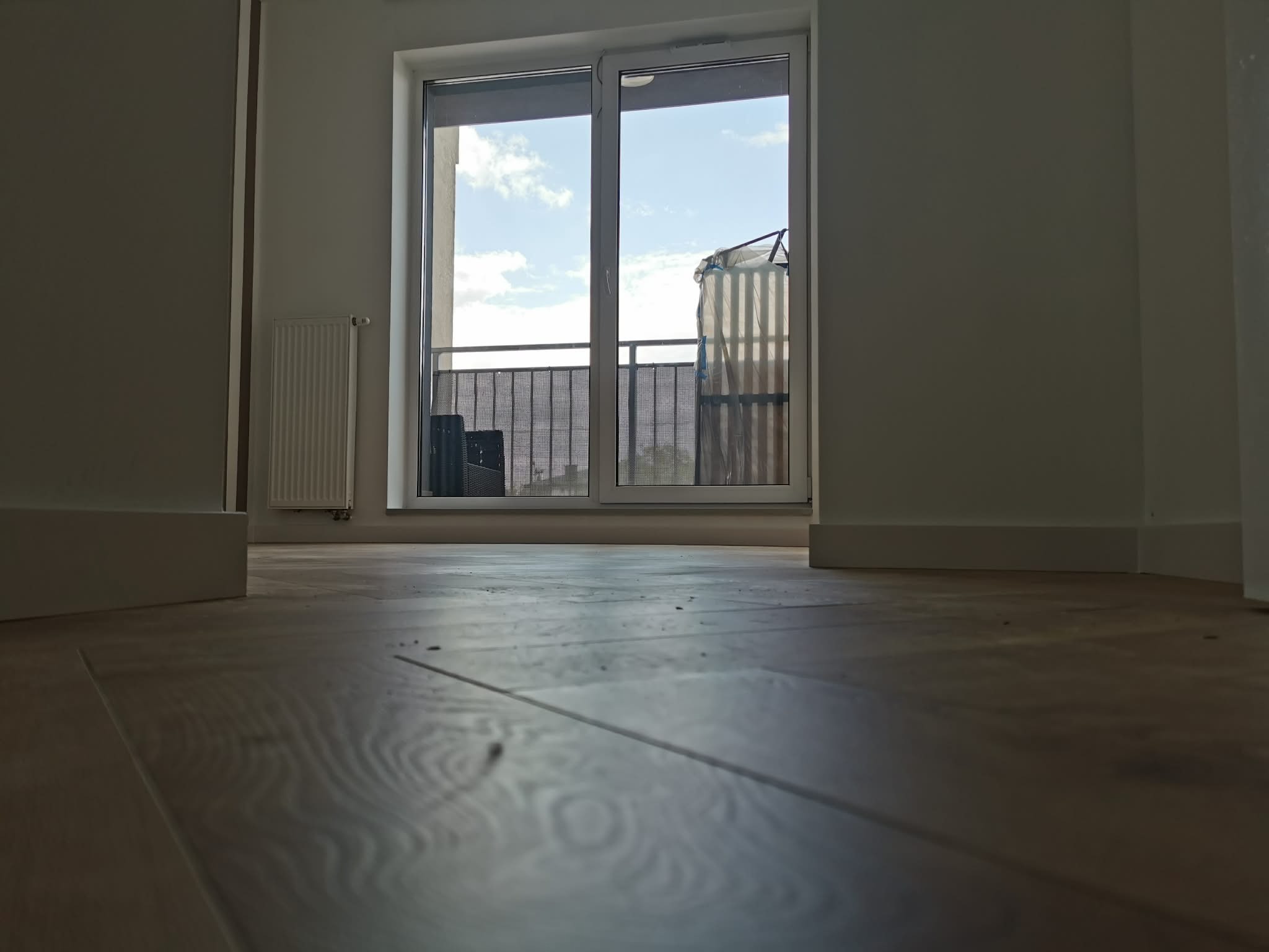 Jasne panele podłogowe w nowym mieszkaniu z widokiem na balkon. Minimalistyczne wnętrze z grzejnikiem i białymi ścianami. Ujęcie z niskiej perspektywy.
