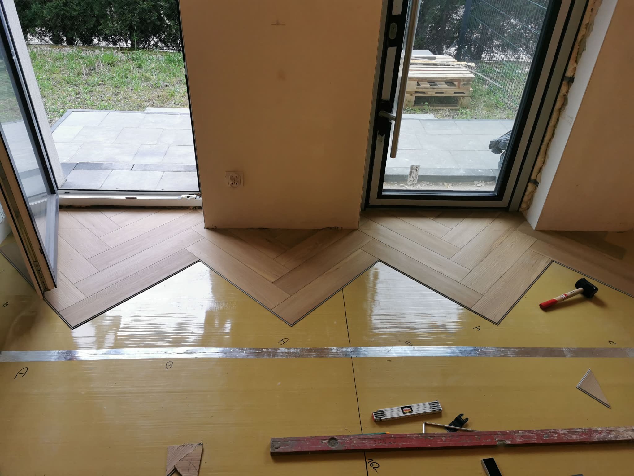 Fragment układania parkietu w jodełkę francuską, widoczne narzędzia i podkład. Częściowo ułożona podłoga w pomieszczeniu z oknami i drzwiami balkonowymi.