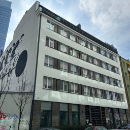 Joanna Pietruk-Fal Property Geek - Elewacja budynku z muralem rowerzystów na tle wieżowca. Widoczne okna z ciemnymi obramowaniami i fragmenty sąsiednich budynków. U dołu graffiti na murze.