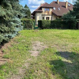 Joanna Pietruk-Fal Property Geek - Dwupiętrowy dom z balkonem, otoczony zielenią i żywopłotem. Widok na zadbany trawnik i drzewa iglaste w słoneczny dzień. Dom z brązowym dachem i jasną elewacją.