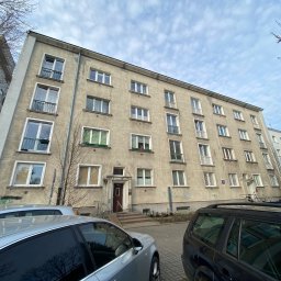 Joanna Pietruk-Fal Property Geek - Szary blok mieszkalny z balkonami i zaparkowanymi samochodami. Widoczne detale architektoniczne i otoczenie budynku. Ujęcie z dołu, pochmurne niebo.