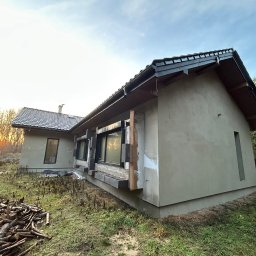 Joanna Pietruk-Fal Property Geek - Niedokończony dom jednorodzinny z szarą elewacją i czarną dachówką, widoczny stos drewna na trawie, w tle zachodzące słońce. Budynek w trakcie prac wykończeniowych.