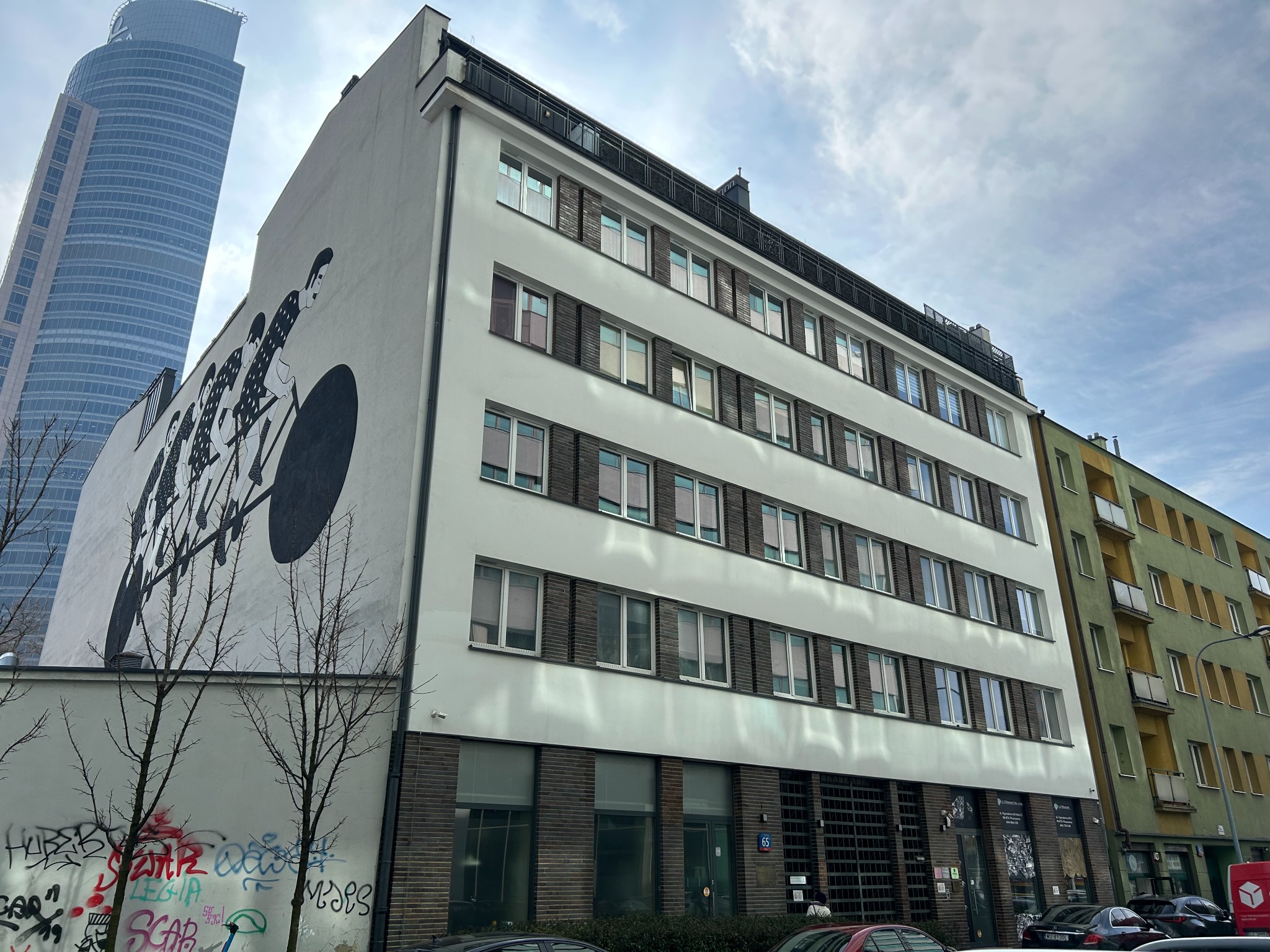 Elewacja budynku z muralem rowerzystów na tle wieżowca. Widoczne okna z ciemnymi obramowaniami i fragmenty sąsiednich budynków. U dołu graffiti na murze.