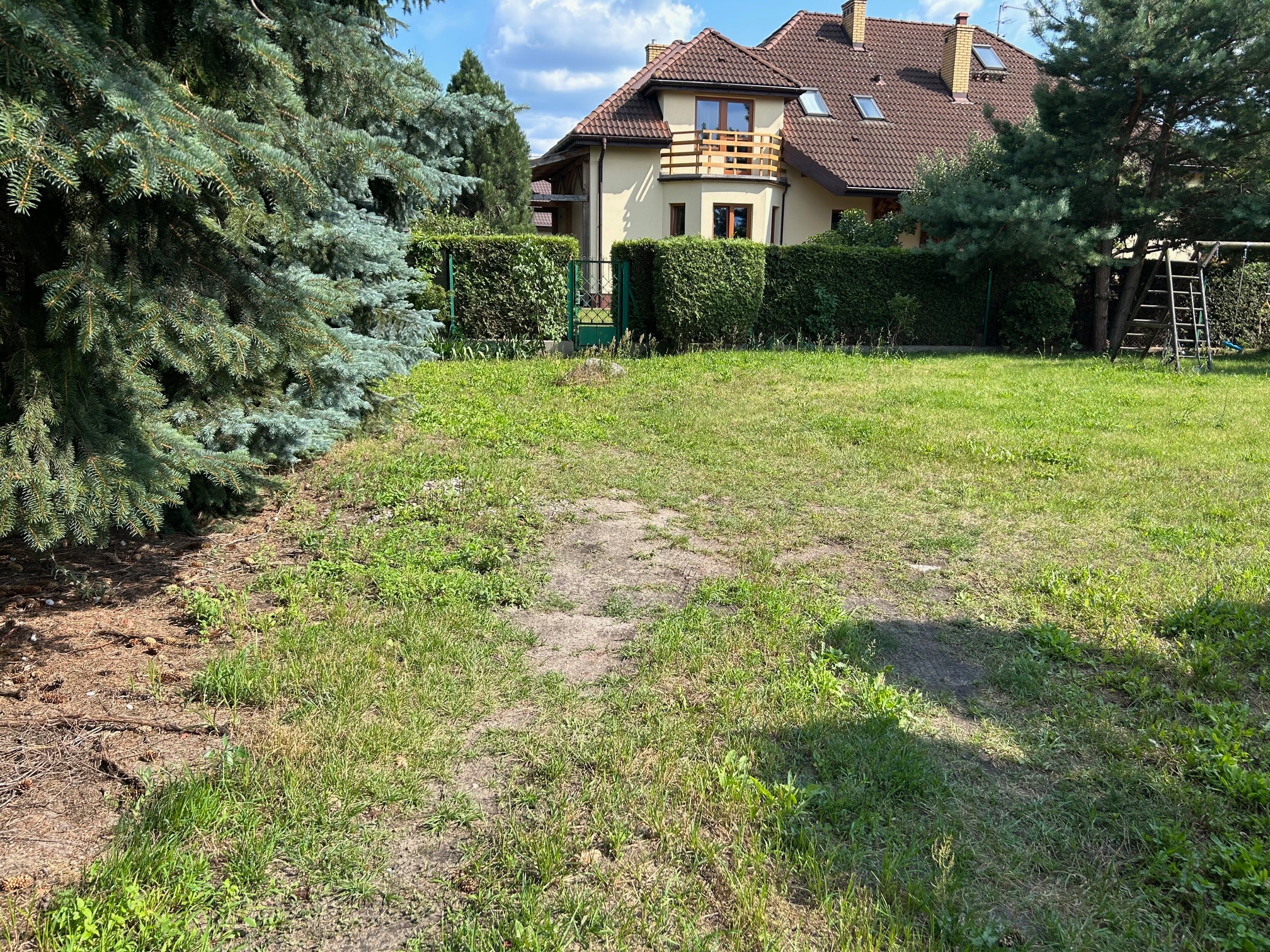 Dwupiętrowy dom z balkonem, otoczony zielenią i żywopłotem. Widok na zadbany trawnik i drzewa iglaste w słoneczny dzień. Dom z brązowym dachem i jasną elewacją.