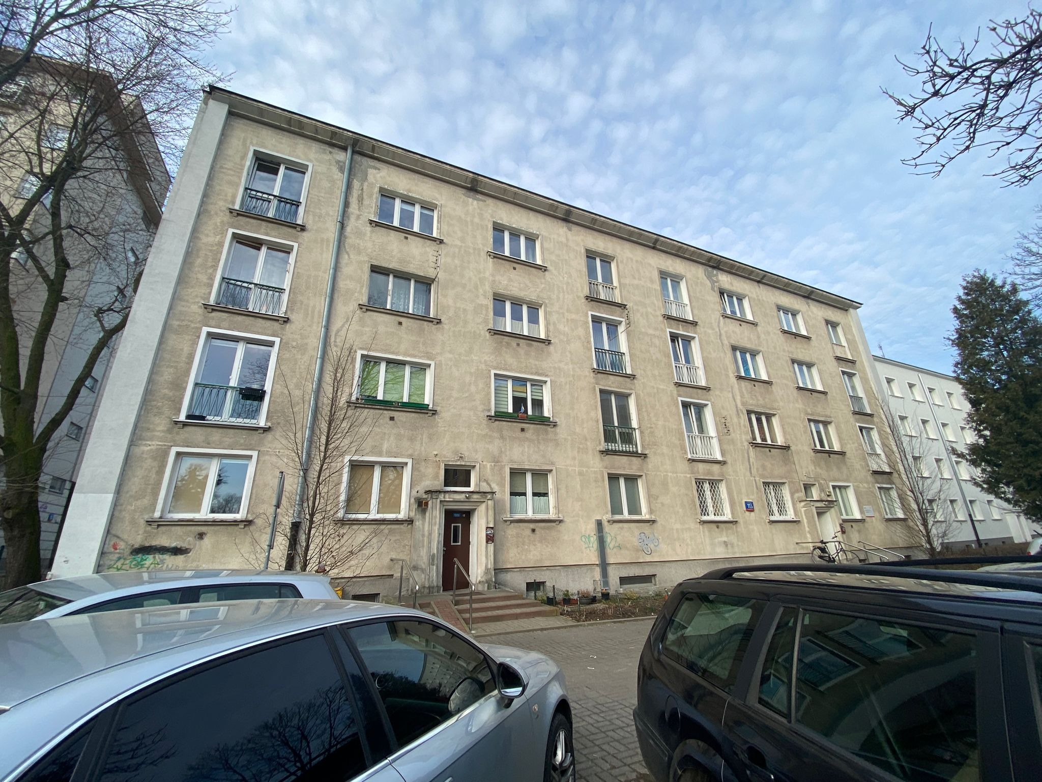 Szary blok mieszkalny z balkonami i zaparkowanymi samochodami. Widoczne detale architektoniczne i otoczenie budynku. Ujęcie z dołu, pochmurne niebo.