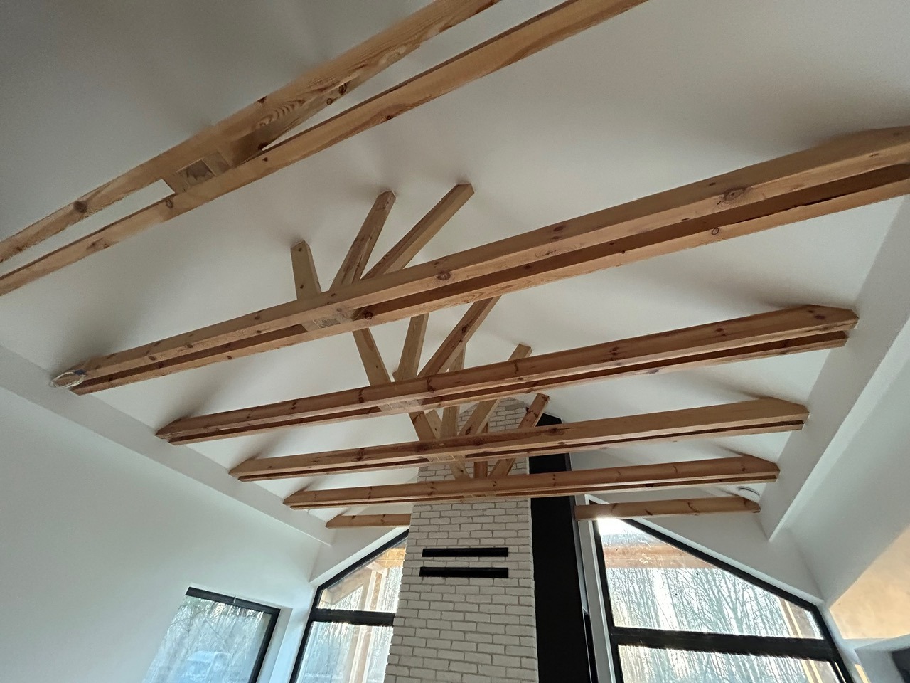 Drewniana konstrukcja stropu z widocznymi belkami i kominem z cegły w nowoczesnym wnętrzu. Widok z dołu, jasne oświetlenie.