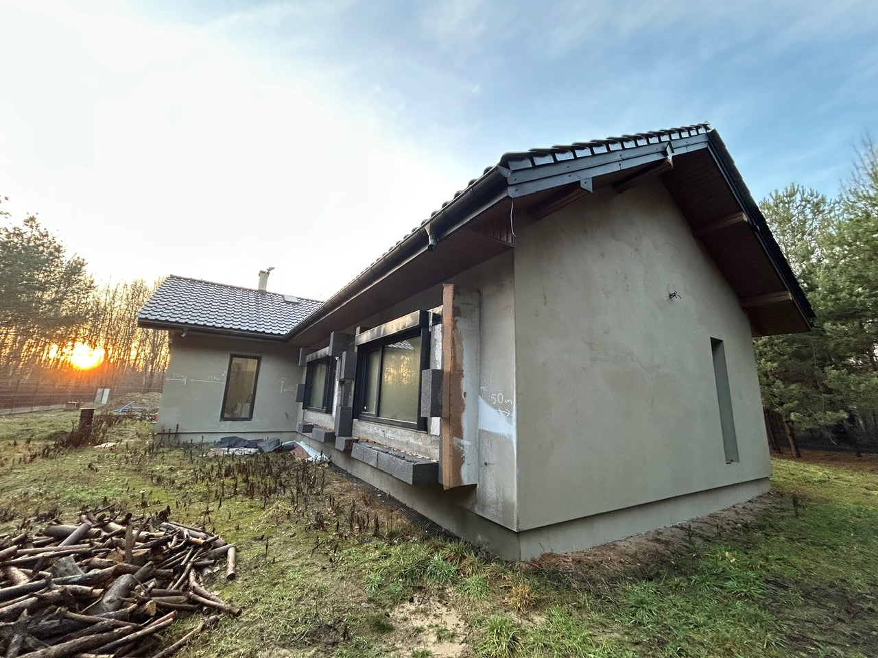 Niedokończony dom jednorodzinny z szarą elewacją i czarną dachówką, widoczny stos drewna na trawie, w tle zachodzące słońce. Budynek w trakcie prac wykończeniowych.