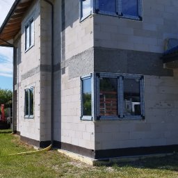 Surowy stan budynku z zamontowanymi oknami PCV, widoczne detale montażu i struktura ścian. Budynek w trakcie budowy, otoczony trawą.