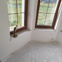 DOBRY MONTAŻ Mariusz Szpakowski - Montaż okien drewnianych z brązowymi ramami w nowym budynku. Na parapecie stoi narzędzie wielofunkcyjne DeWalt. Widok na zewnątrz przez okno.