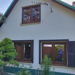 DOBRY MONTAŻ Mariusz Szpakowski - Dom z brązowymi oknami, w których odbija się otoczenie i osoba robiąca zdjęcie. Na ścianie antena. Otwarta okiennica po prawej stronie.