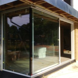 DOBRY MONTAŻ Mariusz Szpakowski - Duże, przesuwne drzwi szklane z białymi ramami w nowym budynku z cegły, widoczne materiały budowlane wewnątrz. Częściowe odbicie osoby w szkle.