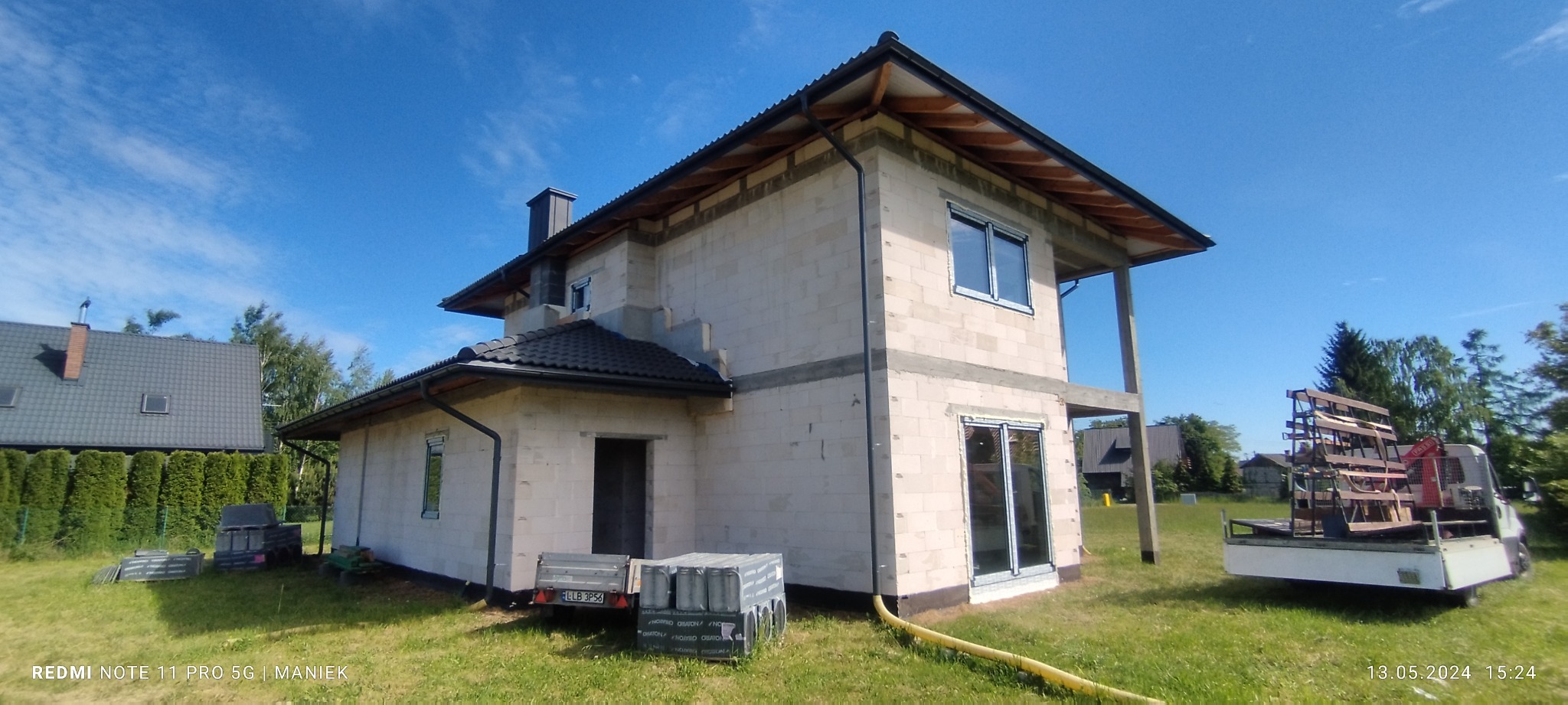 Surowy stan dwukondygnacyjnego domu z białego bloczka, z widocznymi oknami i drzwiami przesuwnymi. Na posesji przyczepka i auto z elementami do montażu.