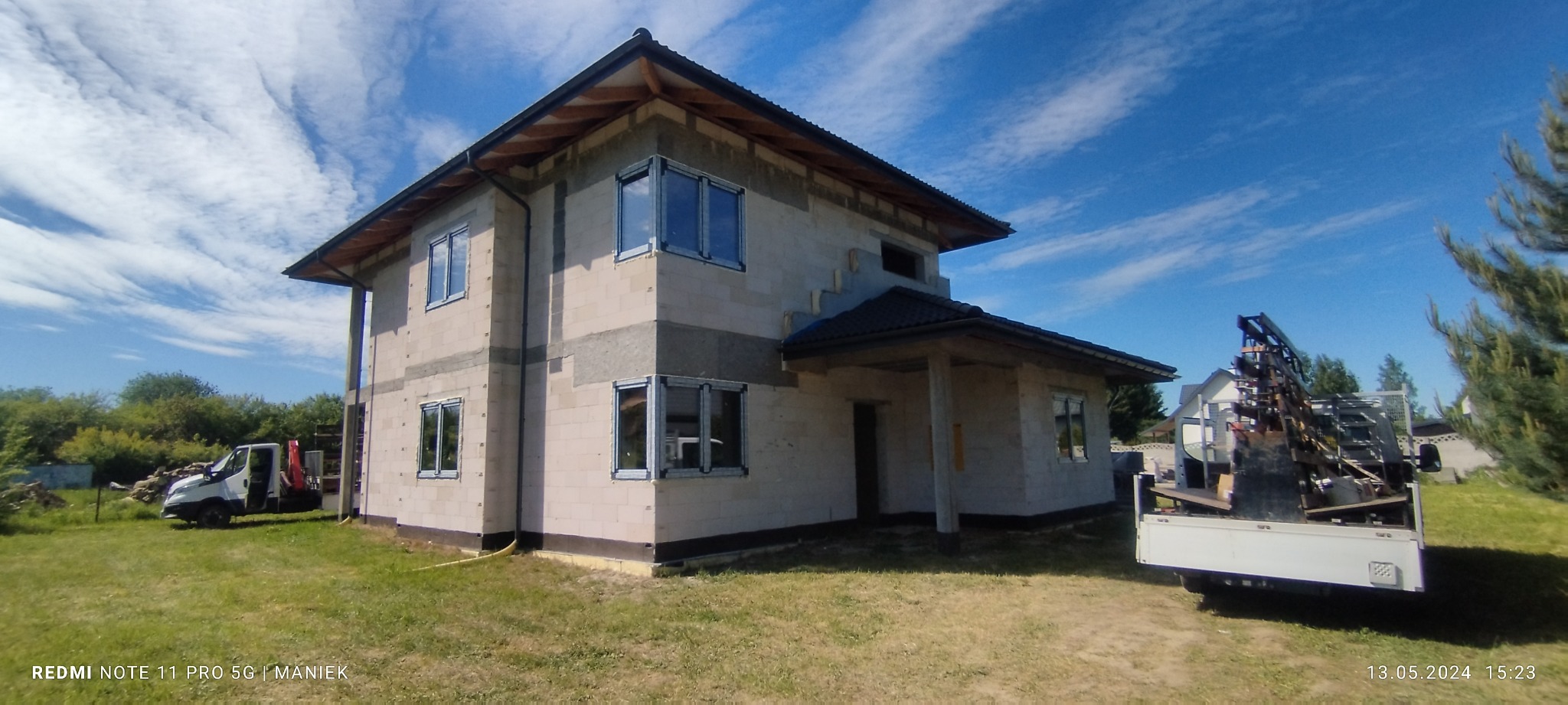 Dom w trakcie budowy z zamontowanymi oknami PCV, widoczny z zewnątrz w słoneczny dzień. Na posesji stoi samochód dostawczy z elementami konstrukcyjnymi.