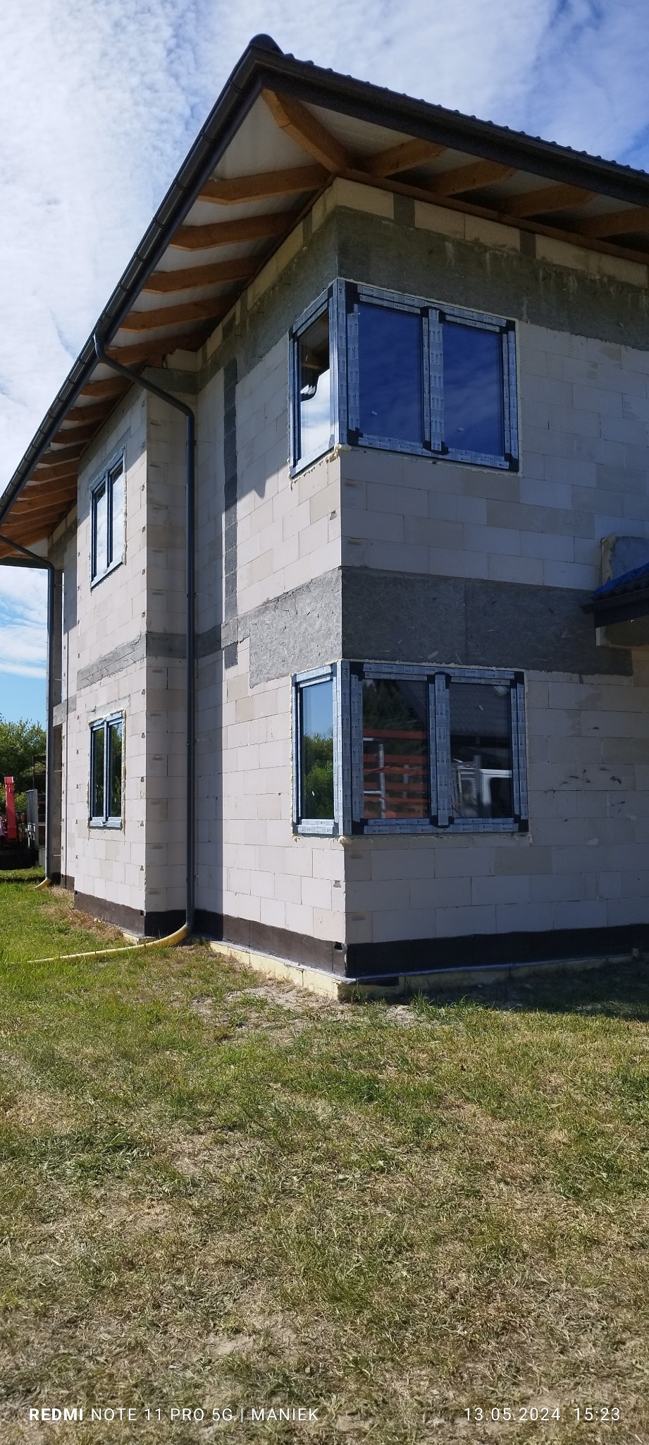 Surowy stan budynku z zamontowanymi oknami PCV, widoczne detale montażu i struktura ścian. Budynek w trakcie budowy, otoczony trawą.